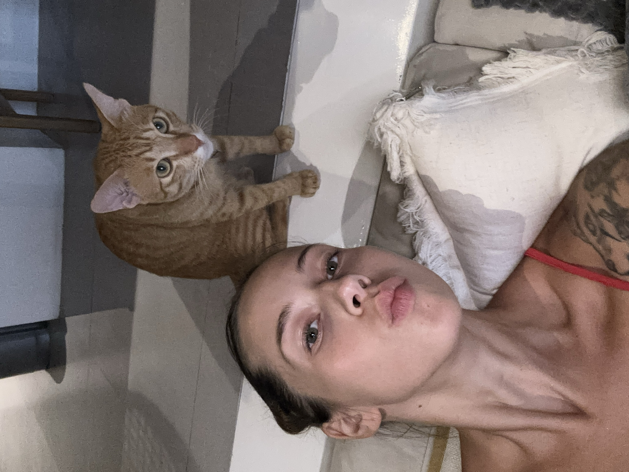 A woman lies supine on a cushioned surface, positioned next to a large, orange tabby cat. The woman's face is angled towards the camera, her lips pursed. A pillow with fringed edges is located to her right. A portion of a table or counter is visible behind the cat. The scene is set indoors. The exact location in Mengwi, Indonesia cannot be determined from the image.