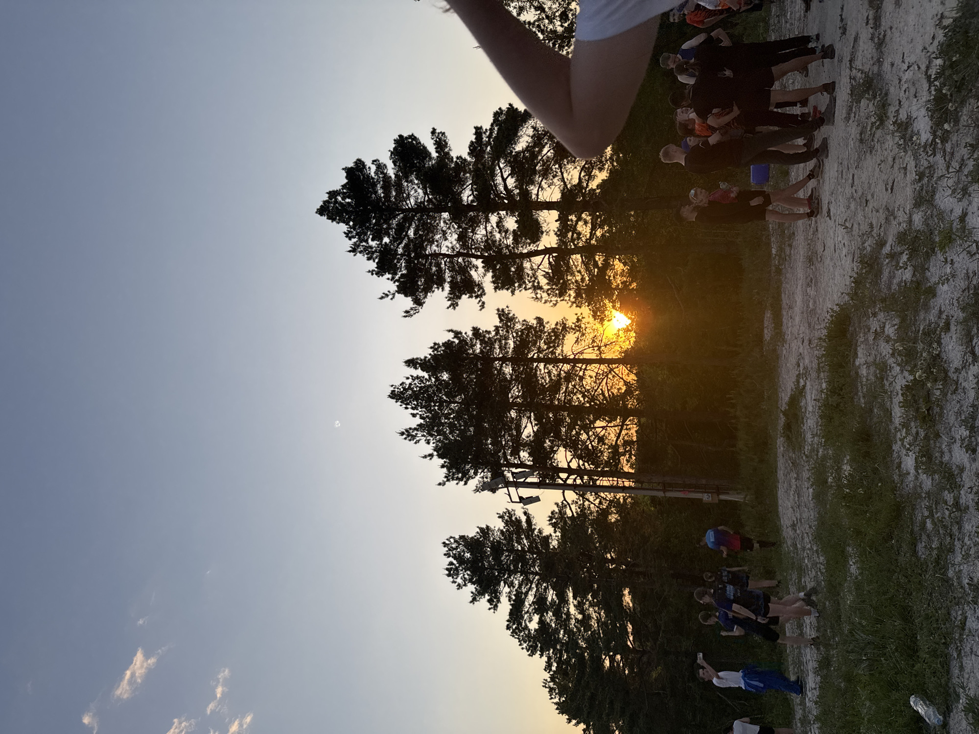The image shows a sunset behind trees, with a group of people standing on a path in Khvalynsk, Russia. A utility pole with lights is visible behind the trees. Some people are looking at their phones.