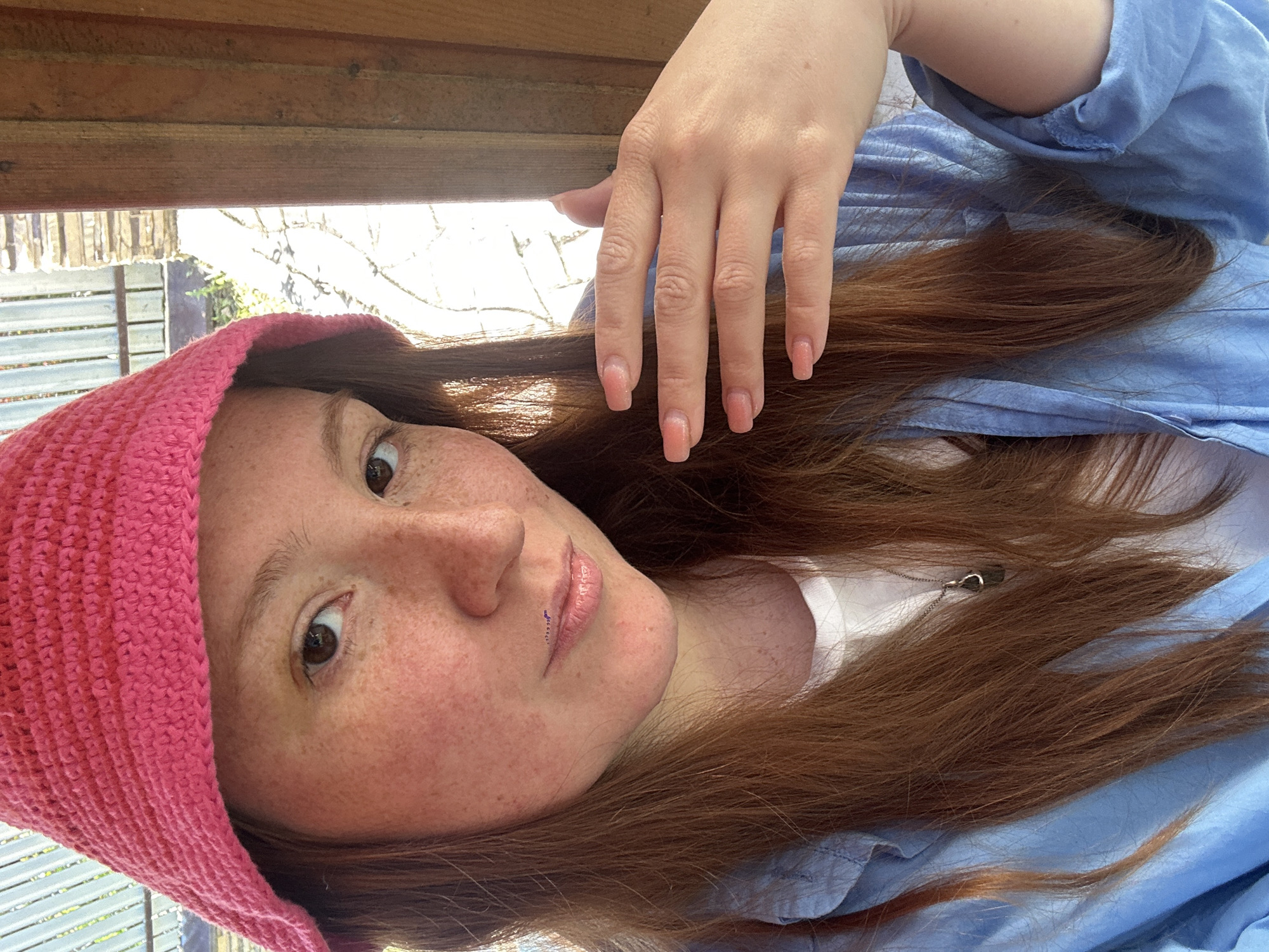 A portrait-style photograph features a young woman with long, reddish-brown hair, fair skin, and numerous freckles across her face. She has brown eyes and a neutral, direct gaze towards the viewer. She is wearing a bright pink, textured knitted hat, a light blue collared shirt, and a white undershirt. A silver necklace with a small pendant is partially visible near her collarbone. A subtle, possibly decorative, mark or piercing is visible above her upper lip, slightly to the left. Her fingernails have light pink polish. Her left hand is visible, resting on a wooden beam or railing that forms part of the background structure, showing the same light pink nail polish.
The woman is positioned outdoors, with bright sunlight and vegetation visible in the background, suggesting a sunny daytime setting. The wooden structure behind and above her head appears to be part of a porch, balcony, or a shaded outdoor area, with glimpses of a lighter-colored fence or building beyond. The scene takes place in Nizhny Novgorod, Russia.