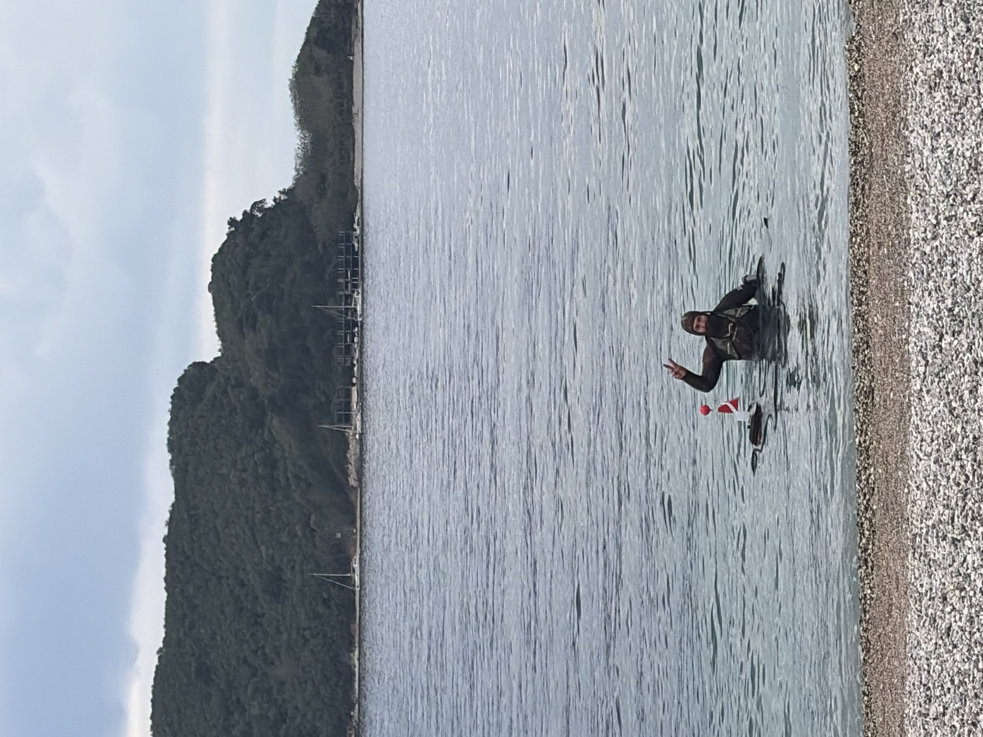 A landscape-oriented photograph shows a man standing in water near the shore, giving a peace sign with his left hand. He appears to be wearing a dark wetsuit or diving gear, including a hood. A small buoy or float with a red and white marker is visible near him in the water.
The background includes a shoreline covered with pebbles, and a large, densely vegetated hill. In the distance, along the base of the hill, there appear to be docks or moorings for boats.
Given the topography and appearance of the landscape, it is likely this photo was taken in or near Budva, Montenegro.