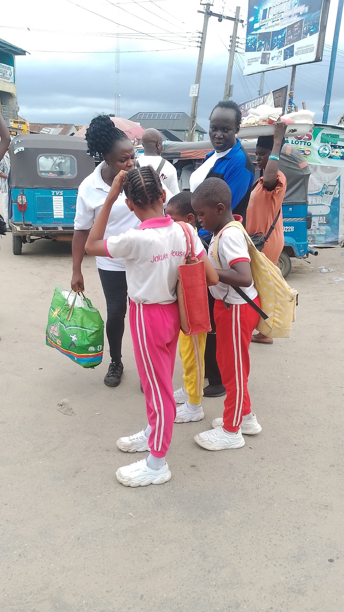 The image depicts a group of people in an urban environment, likely in Ukan, Nigeria, indicated by the presence of a “TVS” branded vehicle commonly used as a public transportation tricycle. In the foreground, there are three young children wearing school uniforms with "Jakuke House" printed on them. Their uniforms consist of white tops with colored trim and matching track pants. They are standing on a paved surface, appearing to be engaged in conversation or listening to an adult nearby. The adult, a woman wearing a white top and black leggings, seems to be attending to the children. In the background, other individuals are visible, including a woman in a blue shirt and another woman in an orange outfit carrying a bag. Behind them, there is a tricycle and an advertisement billboard that includes the words "KINGDOM P". The overall scene appears to be a typical street setting with mixed activity and possibly related to school transportation.