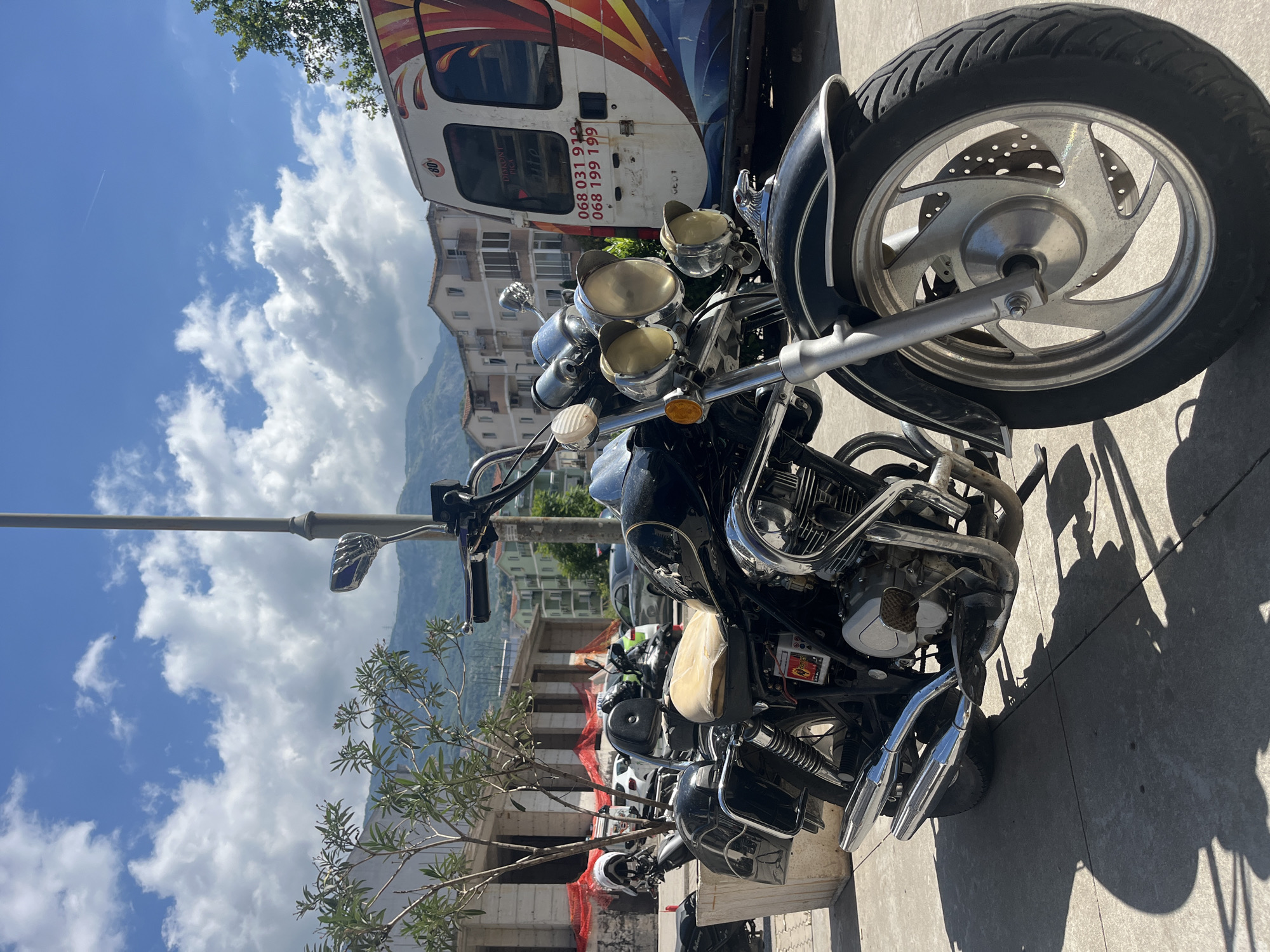 A custom cruiser motorcycle is parked on a paved surface under a bright blue sky with scattered cumulus clouds. The motorcycle, primarily dark in color with extensive chrome accents on its engine, handlebars, and exhaust pipes, features prominent headlights and a light-colored seat. Its front wheel has a distinctive five-spoke design with a visible disk brake. Strong sunlight casts long, dark shadows from the motorcycle onto the ground.
Behind the motorcycle, a white panel van is parked, adorned with colorful abstract graphics in red, orange, yellow, and blue. Visible text on the van includes "DIKONT TRG," phone numbers "068 031 910" and "068 199 199," and a smaller "Lite" logo. A red circular sticker with the number "80" is also present.
In the mid-ground, several multi-story buildings, some appearing residential, are visible. One building on the left shows signs of construction or renovation with orange safety netting. Distant mountains rise in the background under the clear sky. Green foliage from trees is visible, and a street lamp is positioned near the motorcycle's handlebars. A faint airplane contrail can be seen high in the sky. The scene is a daytime exterior shot, likely in a town or city with a mountainous backdrop, consistent with the given location context of Budva, Montenegro.