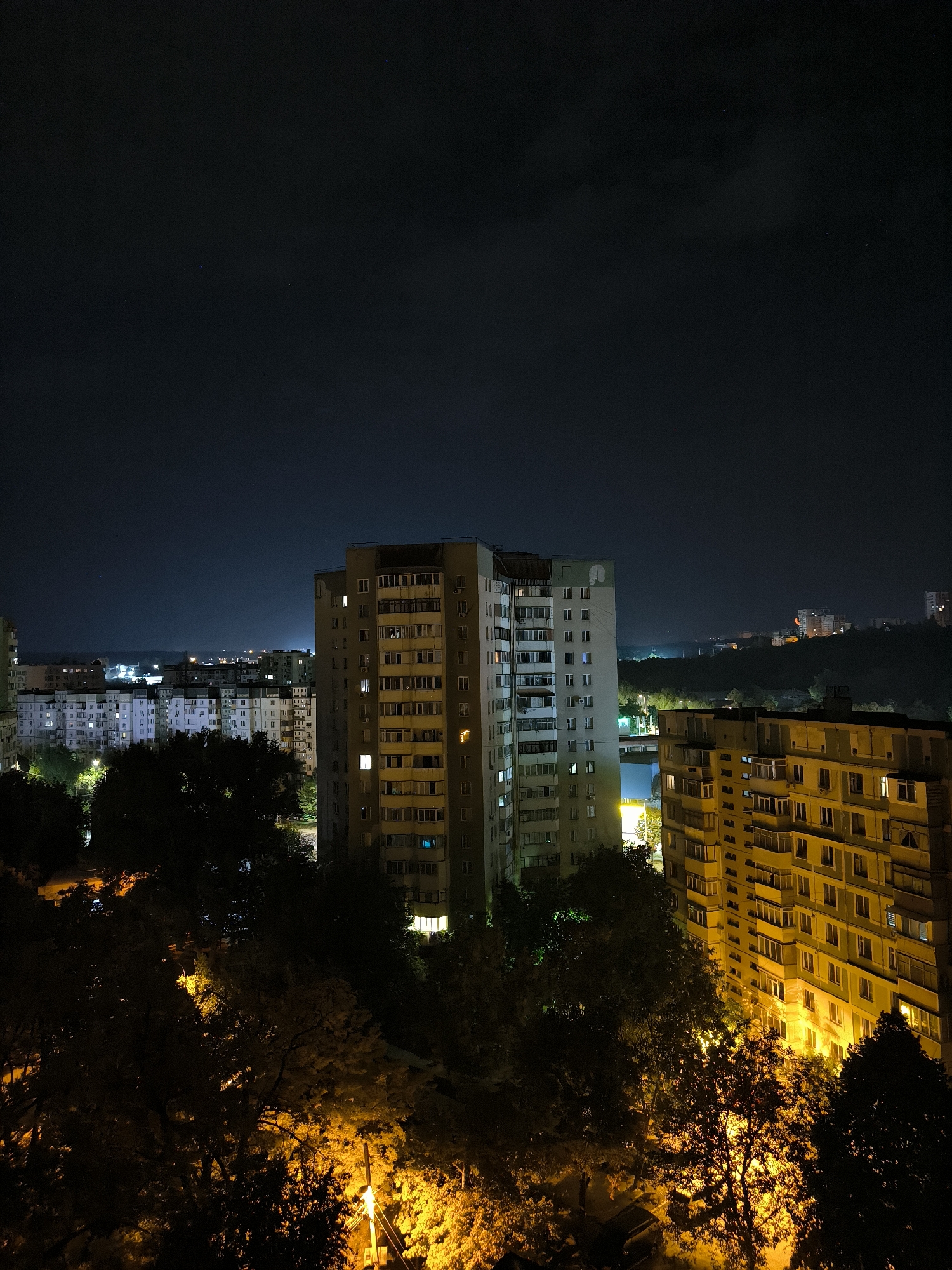 An aerial nighttime photograph captures a view of apartment buildings in Chișinău, Moldova. In the foreground, dark trees with some illuminated foliage from streetlights are visible. Behind the trees, two tall apartment buildings dominate the midground. The building on the left is lighter in color, while the building on the right is yellow and appears to have more windows illuminated. Further in the background, more apartment buildings are visible, stretching into the distance, with some appearing to be on a hillside. The sky above is dark and clear, with a few faint stars visible. The overall impression is of a quiet residential area at night.