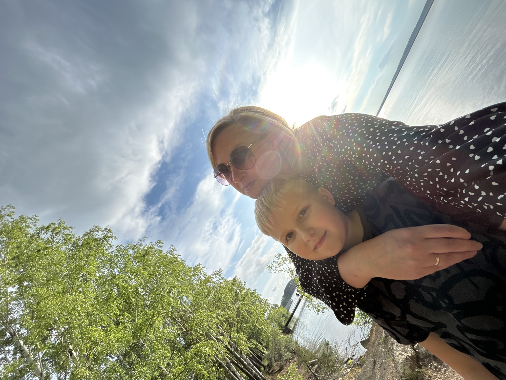 This is a selfie taken outdoors on a sunny day with some clouds in the sky. The primary subjects are a woman and a young boy. The woman is wearing sunglasses and a black dress with white polka dots, and she has her arm around the boy. The boy is wearing a black t-shirt with a gray pattern and is looking towards the camera. He has his arm around the woman's neck. The scene appears to be a natural setting, with a body of water and trees in the background. Some of the trees have light green foliage, suggesting it is spring or summer. The sun is bright and casting lens flares into the image. The overall mood seems happy and affectionate, with the two individuals posing closely together. The location context provided is Chaikovsky, Russia. There is no visible text in the image.