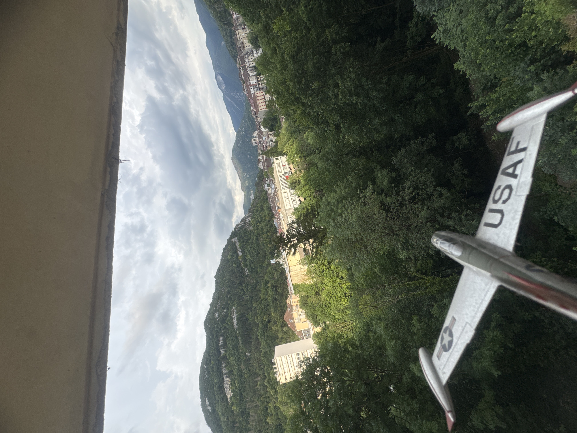 A model airplane, painted silver with red accents and bearing the "USAF" insignia and a star insignia, is positioned in the foreground, angled towards the bottom right of the frame. The airplane's wing displays the letters "USAF" in black.
The background reveals a city nestled in a valley, surrounded by lush, green, tree-covered mountains. The city features a mix of architectural styles, including modern apartment buildings and older, more traditional structures with red-tiled roofs. The sky is overcast with dramatic, cloudy formations, suggesting a potentially stormy or humid day.
The view appears to be from an elevated position, possibly a balcony or window, overlooking the city and its natural surroundings. The location is identified as Saint-Claude, France, a city known for its picturesque setting within the Jura Mountains.