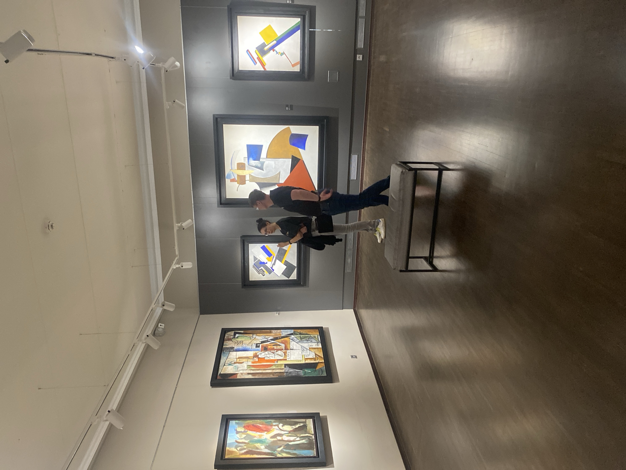 This is a photograph of two people standing in a museum gallery, looking at abstract paintings on the wall. The gallery has dark wooden floors and a white ceiling with track lighting. The paintings are framed and appear to be modern or contemporary art. The two individuals, one woman and one man, are casually dressed. The woman is wearing a black top and grey pants, and the man is wearing a black t-shirt and dark jeans. They are standing in front of a bench placed in the middle of the room, which seems to be part of the museum's visitor amenities. The overall atmosphere suggests a quiet and contemplative setting for art appreciation. The location is not specified in the image itself, however, a search of the paintings suggests this is the Yekaterinburg Museum of Fine Arts.