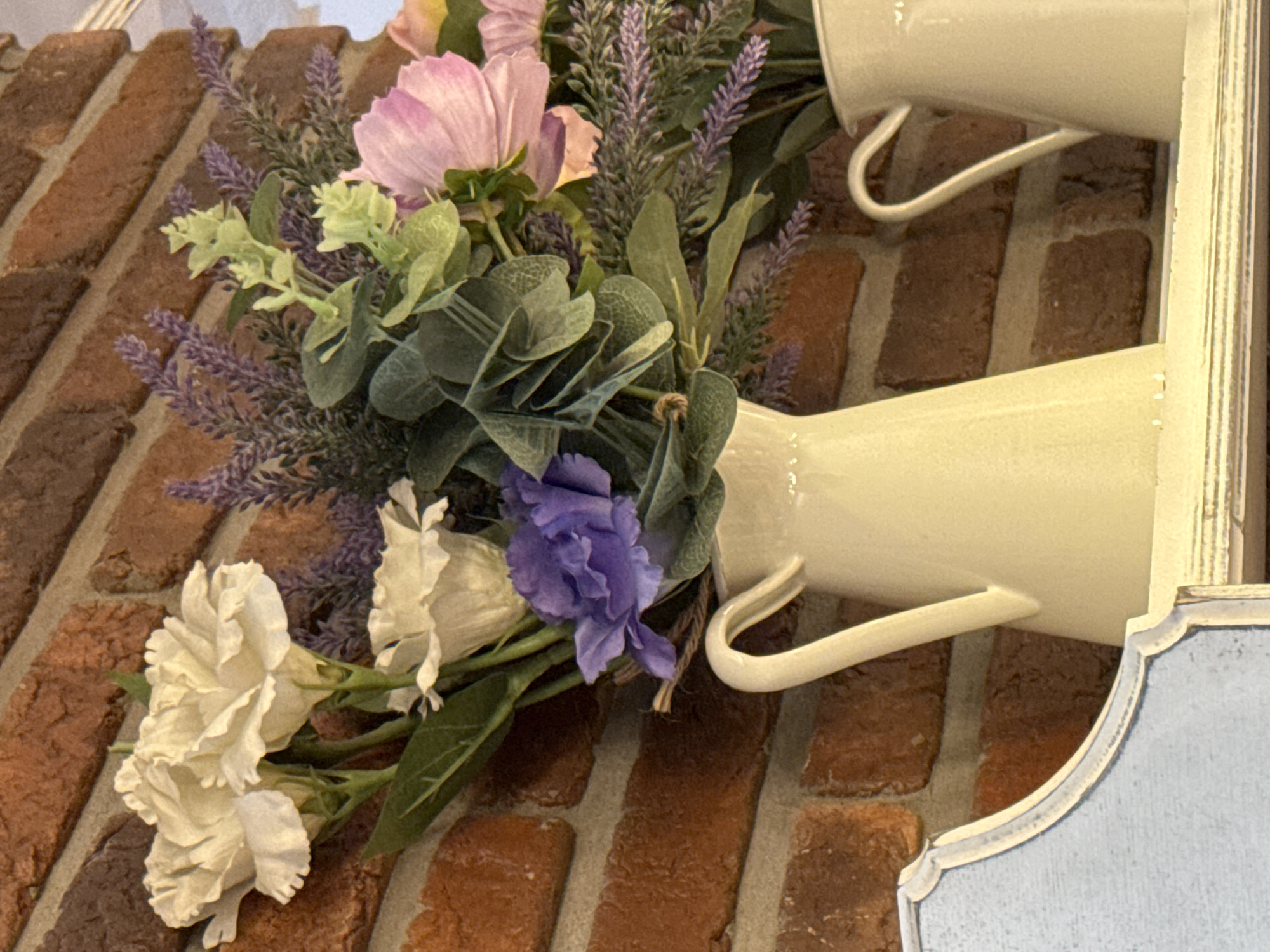 A close-up shot captures a rustic arrangement of artificial flowers in cream-colored pitchers, set against a textured red brick wall. The bouquet features a mix of white, purple, and pale pink flowers, interspersed with green foliage and sprigs of lavender. The pitchers have a slightly worn, enamel finish and graceful handles. The image is framed on the left by a light blue decorative architectural element, possibly part of a shelf or furniture piece. The overall impression is one of quaint, decorative indoor styling. The image does not contain any information that would allow for the determination of its location in Vsevolozhsk, Russia.
