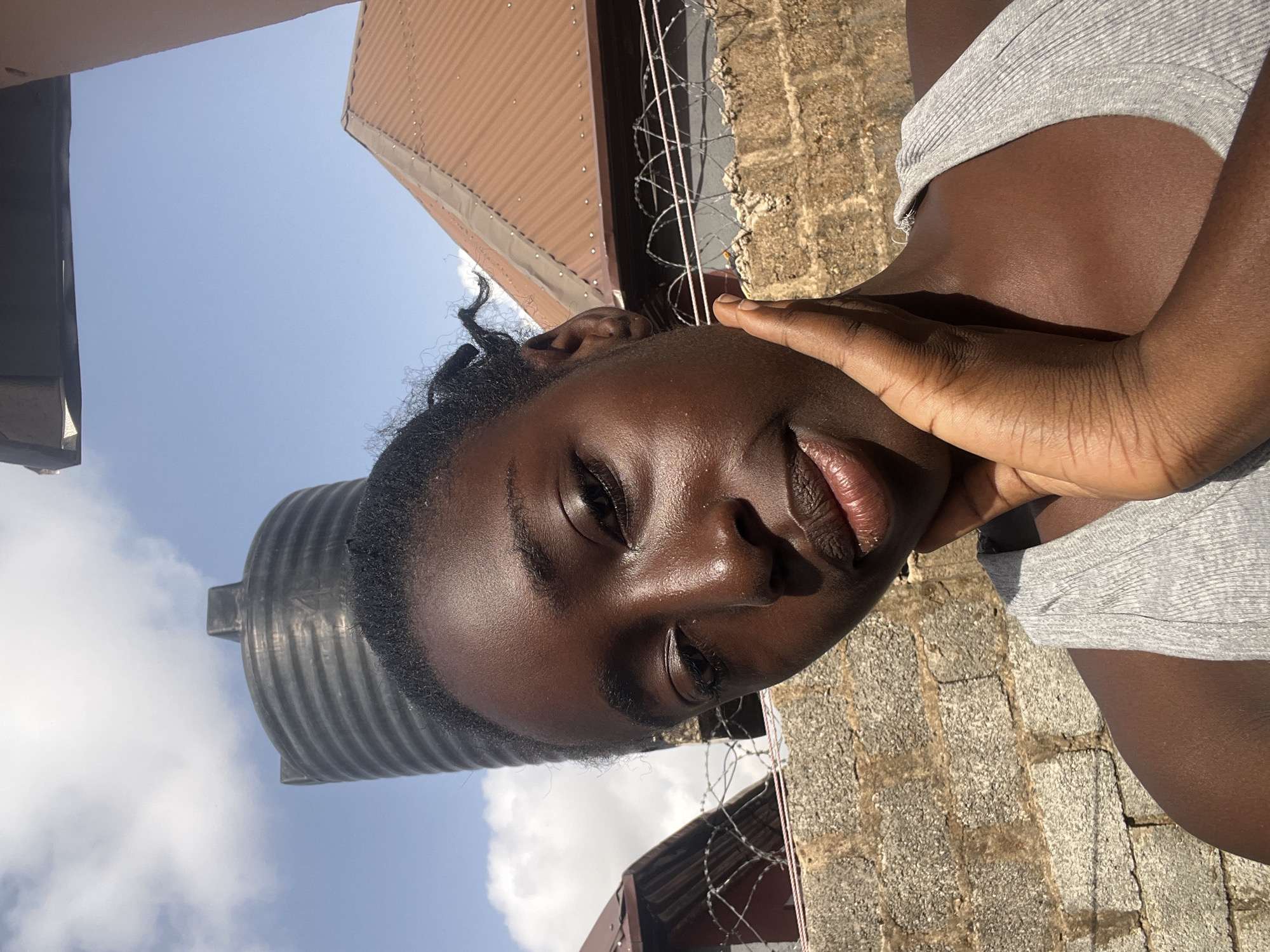 A close-up, low-angle shot of a dark-skinned young woman. She is holding her hand to her cheek, smiling softly at the camera. She is wearing a gray shirt. The background features a blue sky with clouds, a large black water tank, buildings with brown corrugated iron roofs, barbed wire, and a brick wall. The scene appears to be outdoors, possibly in a residential area of Abuja, Nigeria. No specific location within Abuja can be determined.