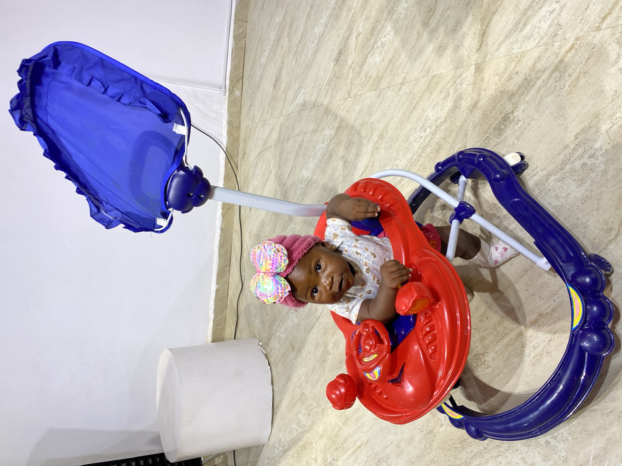 A toddler sits in a red and blue walker with a blue canopy. The toddler is wearing a pink knitted hat with a colorful bow on top, a white shirt with a subtle pattern, and pink socks with white hearts. The walker has a red tray with a steering wheel and toy buttons. The background consists of a light-colored tiled floor and a white wall. A white cylindrical object is visible on the left side of the frame. The scene appears to be indoors, in a home environment, potentially in Gura Top, Nigeria.