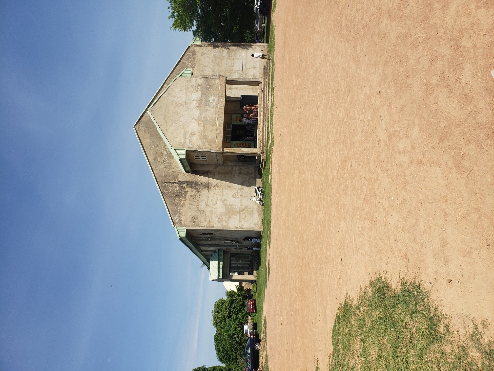 This image depicts an outdoor scene in Narayi, Nigeria, dominated by a large, weathered concrete building under a clear blue sky.
The central subject is a two-story building constructed of light grey, possibly unpainted, concrete, exhibiting signs of wear and age on its surfaces. It features a prominent gabled roof with green-colored eaves and fascias. The main entrance is recessed, fronted by a set of steps, and supported by two square pillars. A group of approximately five to seven individuals, some dressed in patterned attire, are visible on these steps and within the entranceway, suggesting entry or exit from the building. To the left side of the building's main entrance, two individuals, possibly children or adolescents, stand near the wall, adjacent to a stack of wooden planks. A white motorcycle is parked on a patch of grass to the immediate right of the building. Further right and in the background, a lone male in light-colored clothing is standing with his back to the camera, facing the building.
The foreground consists of a vast expanse of reddish-brown, compacted earth or sand, showing subtle tire tracks and footprints. A patch of green grass is visible in the lower-left corner, casting a shadow that extends onto the dirt. To the left of the building, several vehicles are parked on a grassy verge: a black sedan, a white SUV, and a red sedan. Beyond these vehicles and behind the building, dense green foliage and trees are visible, extending to the background on both sides of the structure. The sky is bright, clear blue, with no discernible clouds. A faint, dark, elongated object (possibly a distant aircraft or bird) is visible high in the sky, slightly to the left of the image center. A subtle crescent moon is also visible in the upper-left quadrant of the sky. The scene is illuminated by strong daylight, casting shadows consistent with a high sun angle.