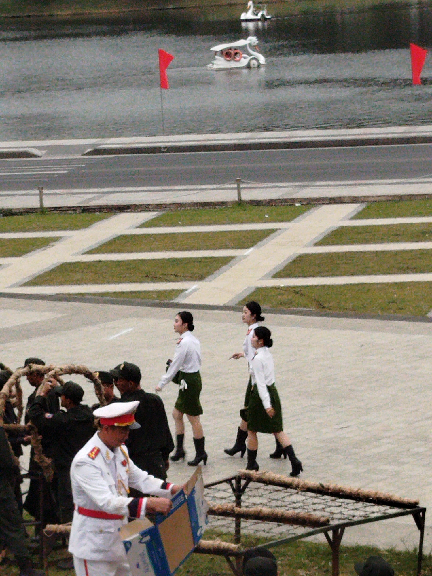The image depicts a scene outdoors, likely in Da Lat, Vietnam, with a body of water in the background featuring two swan-shaped paddle boats. A red flag is planted in the water. In the foreground, a group of people are gathered. A man in a white military uniform with red accents and a red sash is examining a cardboard box. Behind him, several individuals in dark uniforms and hats are visible, some appearing to be carrying a large, circular, rope-like structure. Further in the background, two women in white shirts and green skirts are walking. The overall atmosphere suggests a formal gathering or event, possibly a parade or ceremony, given the uniforms and the organized presentation. The lighting suggests it might be daytime, though the weather appears overcast. No discernible text is visible in the image.