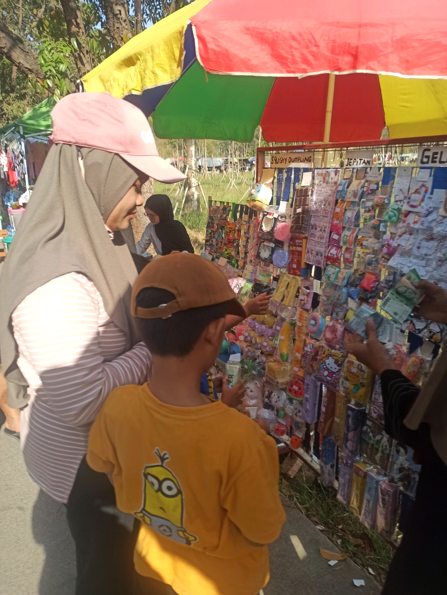 In Telagamurni, Indonesia, a street stall displays an assortment of small toys and accessories under a colorful striped umbrella. A woman wearing a grey hijab and a pink baseball cap stands to the left, facing a boy in a yellow t-shirt with a Minion graphic. The boy is looking at the stall, and his hand is reaching towards it. A woman's hands are visible on the right, holding Indonesian currency, seemingly in the process of making a purchase. The stall is laden with items packaged in plastic, including what appear to be keychains, small toys, and novelty items. The ground is paved, and grass and trees are visible in the background, suggesting an outdoor market setting.