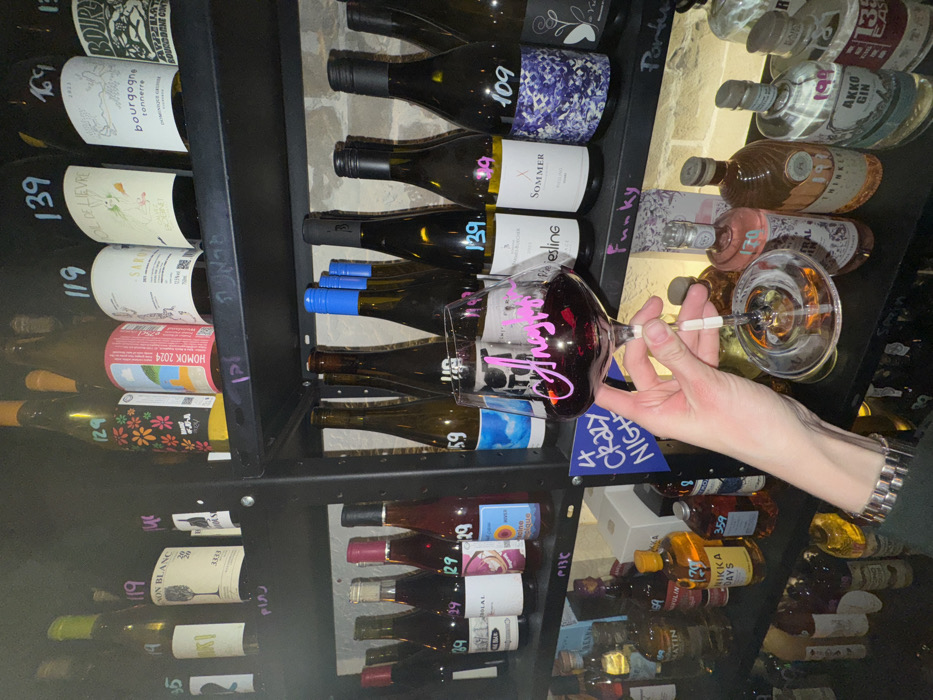 A tilted close-up shot displays shelves filled with various alcoholic beverages. On the left shelves are assorted wine bottles labeled with price tags like "139," "119," and "129." The right shelves feature gin and other liquor bottles. A hand holds a wine glass filled with red wine, with some of the liquid spilled down the side. A small blue sign reads "CRAZY NIGHT." The entire scene is dimly lit, giving it a bar or restaurant atmosphere.