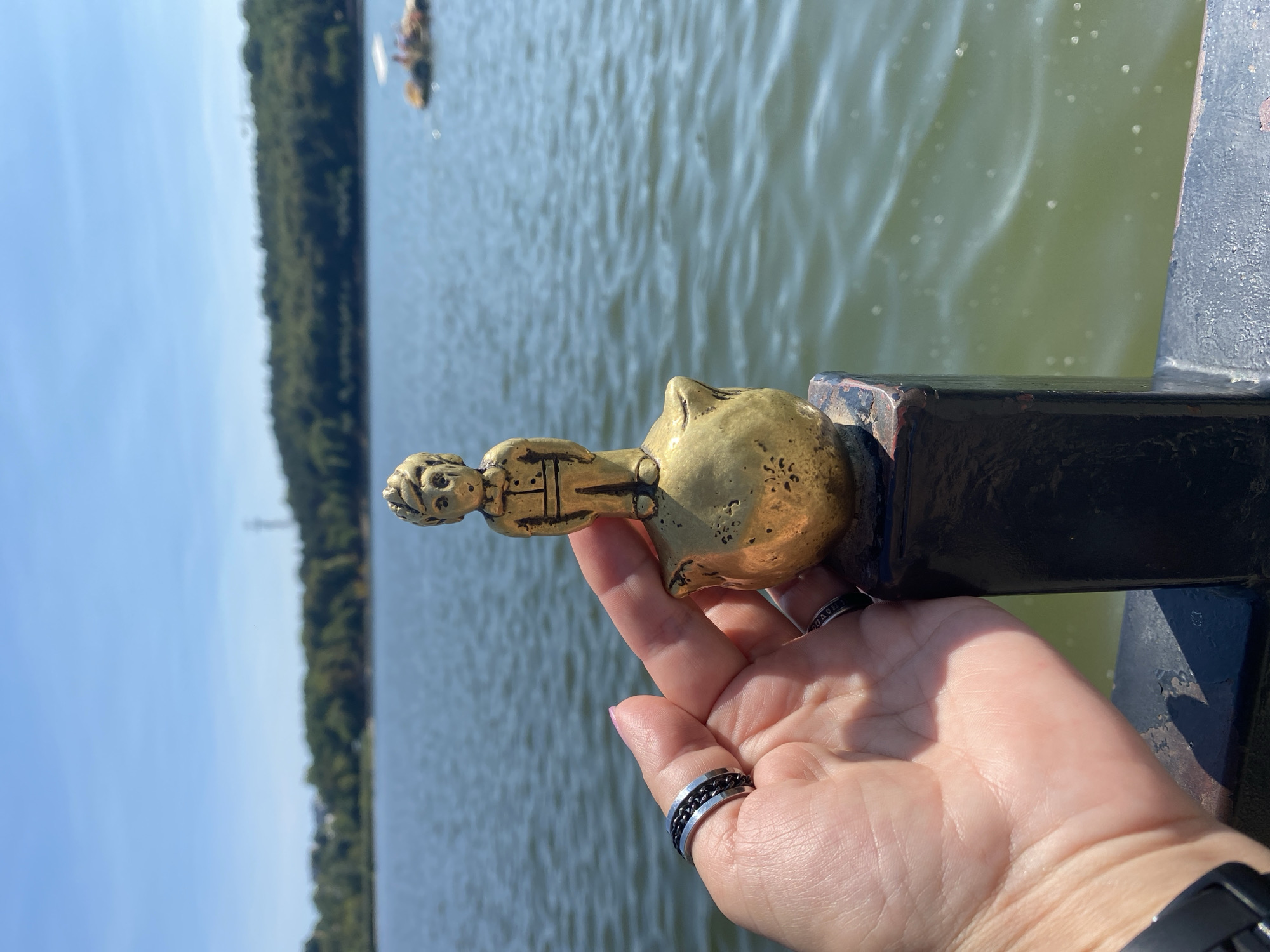 In the foreground, a human hand with two rings on one finger supports a small, golden figurine of a boy standing on a round object, which is affixed to a dark, textured post. The figurine is detailed with a suit and an expressive face. The hand and figurine are positioned over a body of water with gentle ripples under a clear blue sky. In the background, a tree-lined shore is visible, with a distant hill. A small raft with people on it floats on the water. The scene appears to be outdoors, likely at a lake or reservoir. The specific location within Chișinău, Moldova, is not identifiable from the image alone.