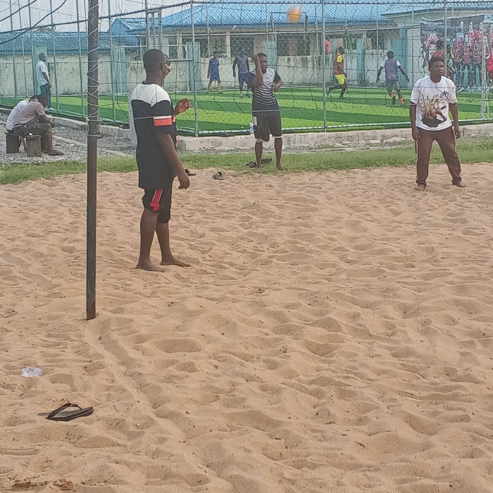 The image displays an outdoor multi-sport recreational area in Koko, Nigeria. In the foreground, a light brown, uneven sand court occupies the lower two-thirds of the frame. A single dark flip-flop and a small piece of white debris are visible on the sand in the lower left. A metal pole, likely part of a net support, stands vertically on the left.
Three adult males are on the sand court, engaged in what appears to be beach volleyball. The man on the left wears a black shirt with white and orange horizontal stripes and black shorts with red trim; he is barefoot and faces right. A second man, positioned centrally to the right, wears a black and white striped short-sleeved shirt and black shorts; he is also barefoot, with his right arm raised towards a yellow ball suspended in the air. A third man on the far right wears a white t-shirt with a graphic design and brown pants, standing barefoot with his feet apart.
Behind the sand court, a wire fence separates it from a green artificial turf field. Several individuals are visible on this turf field, participating in a football (soccer) match, wearing various colored jerseys. In the background, a low building with a light blue corrugated metal roof is visible. On the far left, beyond the sand court, another man is seated on a block, observing the scene.