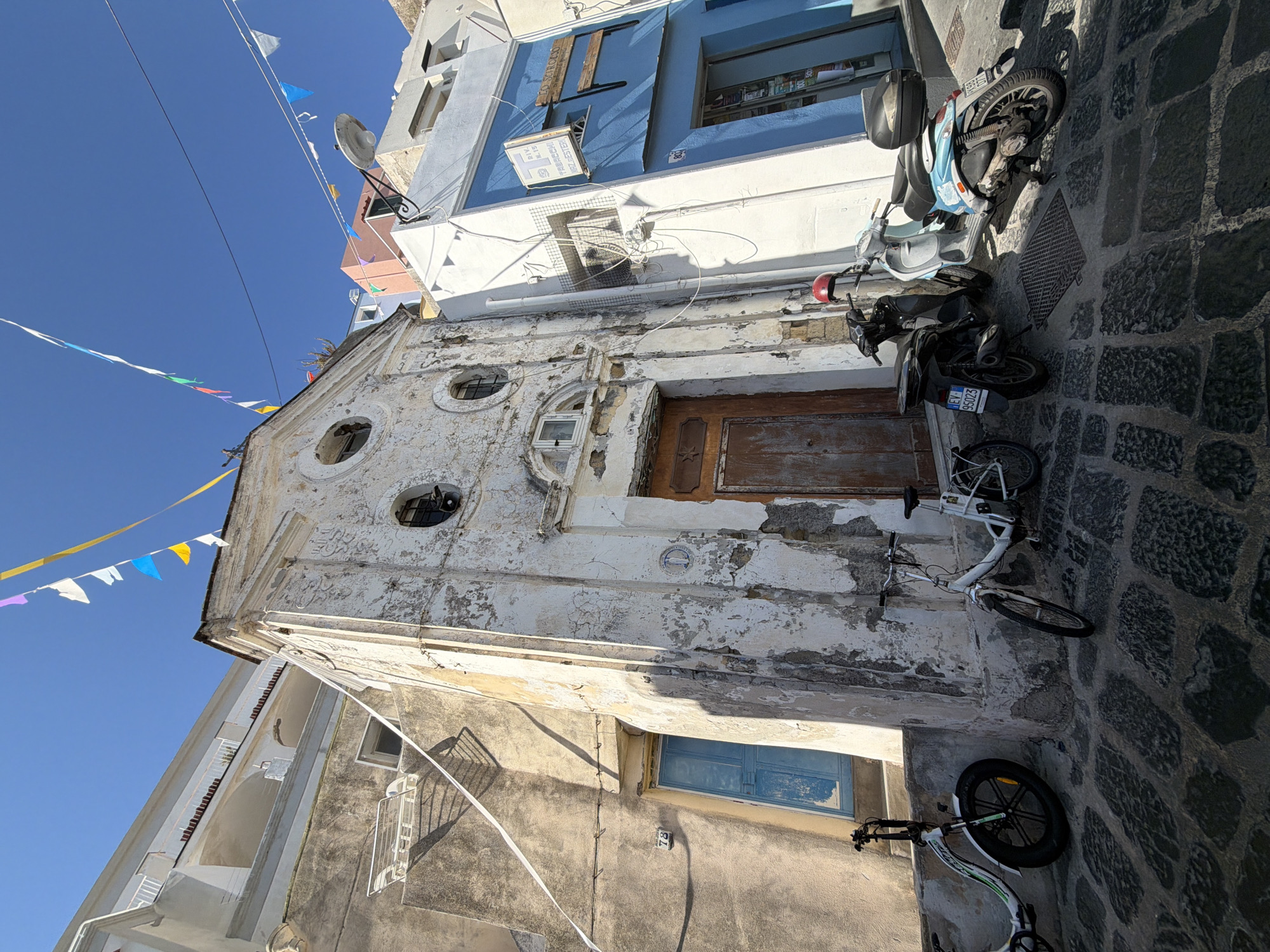 The image depicts a narrow street in Procida, Italy. The street is paved with dark cobblestones. A weathered building with a light-colored facade and a prominent wooden door stands on one side of the street. The building features circular windows and a small window above the door. The facade exhibits signs of age and disrepair, with peeling paint and visible wear. Parked along the street are two scooters and two bicycles, partially obscuring the building's base. Overhead, colorful pennants are strung across the street, adding a festive touch to the scene. Adjacent buildings with blue and white facades rise above the primary building, contributing to the typical architecture of Procida. The sky is clear and blue, suggesting a sunny day.