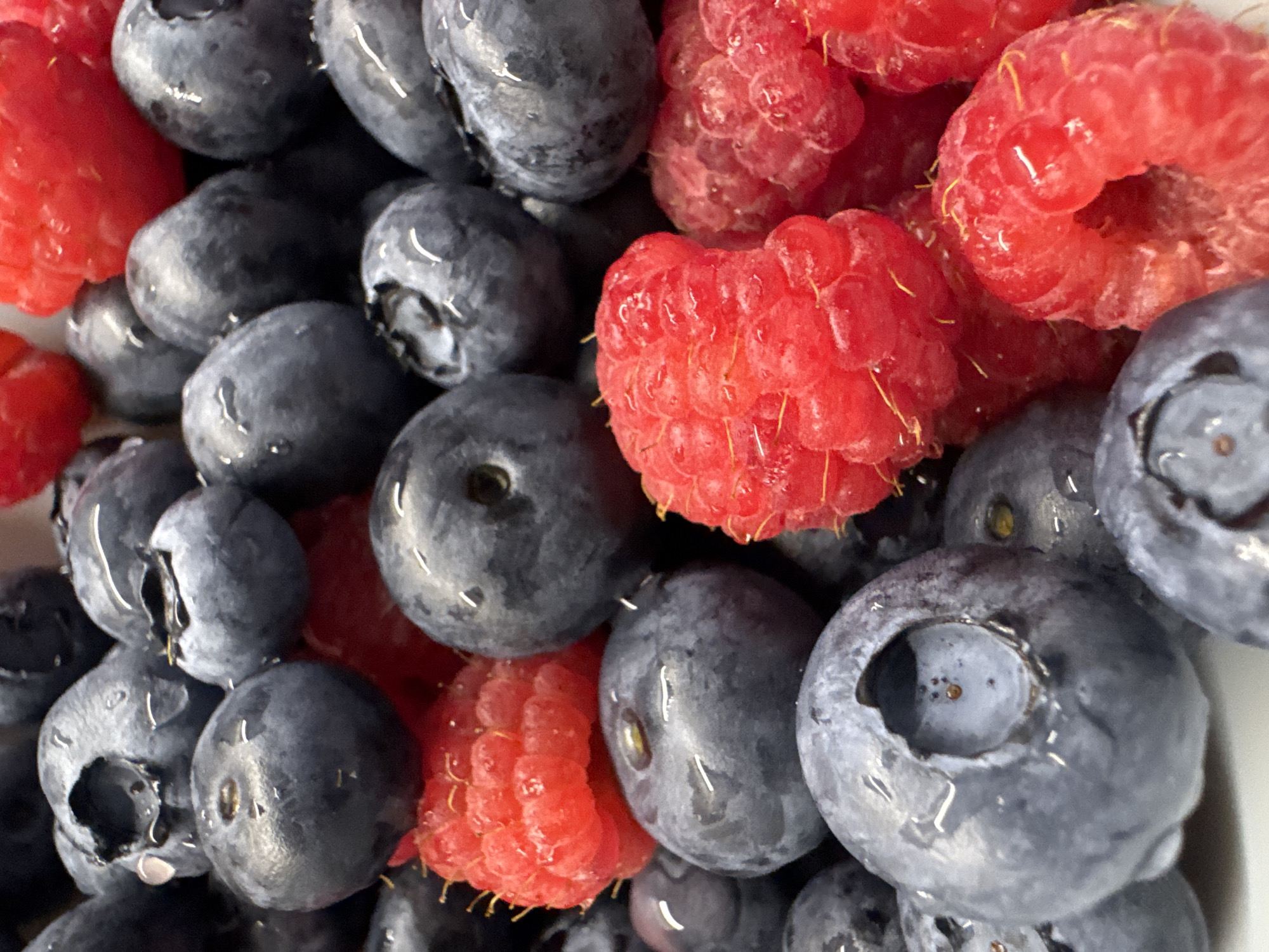 A close-up, top-down shot shows a pile of fresh blueberries and raspberries. The berries are wet, with small water droplets clinging to their surfaces, suggesting they may have been washed or are naturally moist. The blueberries are a deep blue-purple color and appear plump, with some showing the characteristic "flower" at the stem end. The raspberries are a vibrant red, with their segmented drupelets clearly visible. The lighting is bright and even, highlighting the textures and colors of the fruit. The background is dark and out of focus, ensuring the berries are the sole focus of the image. There is no discernible text, time of day, weather, or location cues beyond the fruit itself. The overall impression is one of freshness and natural abundance.