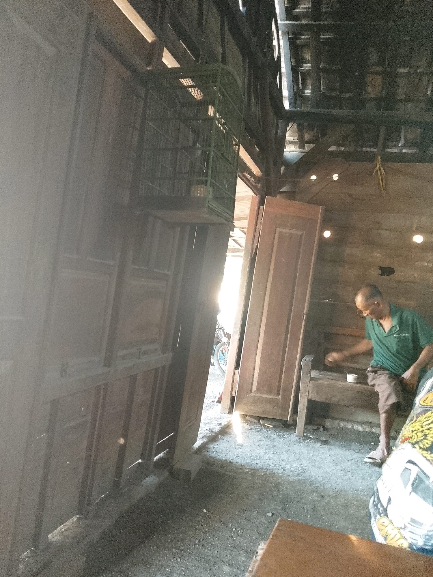 The image depicts an indoor scene, possibly a workshop or storage area, in Wadang, Indonesia. The environment appears rustic, with wooden structures and a gravel floor. In the foreground, a man wearing a green polo shirt and brown shorts is seated on a wooden bench. He is holding a small white cup and appears to be engaged in some activity, possibly related to the objects on the bench. To the left, several large wooden doors are stacked vertically. Above the doors, a green birdcage is visible. The background shows a wooden wall with visible ceiling beams and light fixtures. Sunlight streams in from the left, creating bright highlights and shadows. The overall impression is of a functional, albeit somewhat unorganized, workspace.