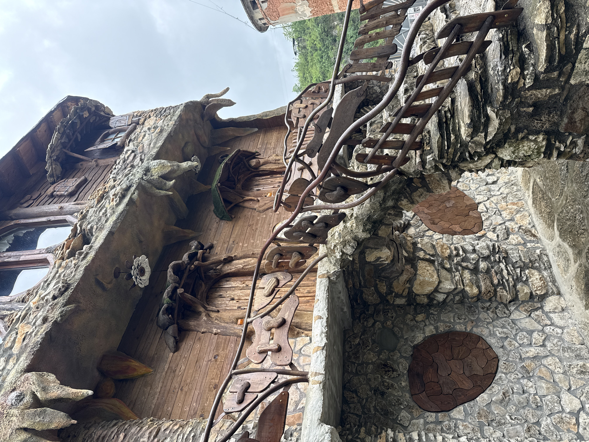 The image shows the unconventional architecture of the building known as the "Stone House", located in Lukovit, Bulgaria. The building features a wooden facade with stone-like decorations and unique wooden sculptures. A wooden staircase with metal handrails is attached to a wall built with various sizes of stones. Two large, circular wooden inlays are visible on the stone wall. The sky is overcast.