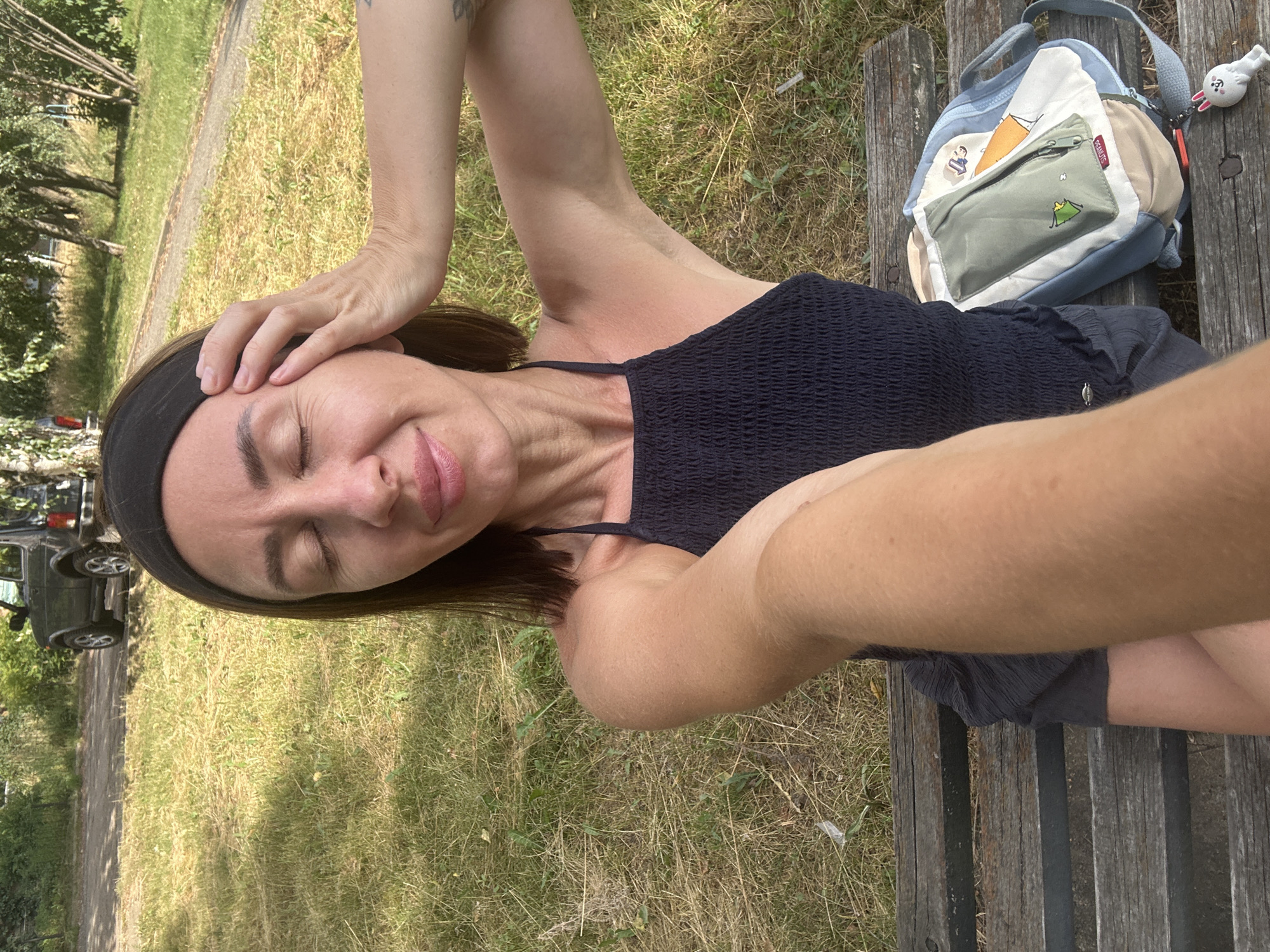 A woman is sitting on a wooden park bench in Votkinsk, Russia, with her eyes closed and her hand on her temple, appearing relaxed. She is wearing a black, smocked halter top and dark shorts. A light blue backpack with a green pencil case and a white rabbit keychain rests on the bench next to her. Behind her, a dark-colored SUV is parked on a dirt road, with a grassy area and trees surrounding the scene. The sun is shining, casting shadows on the ground.