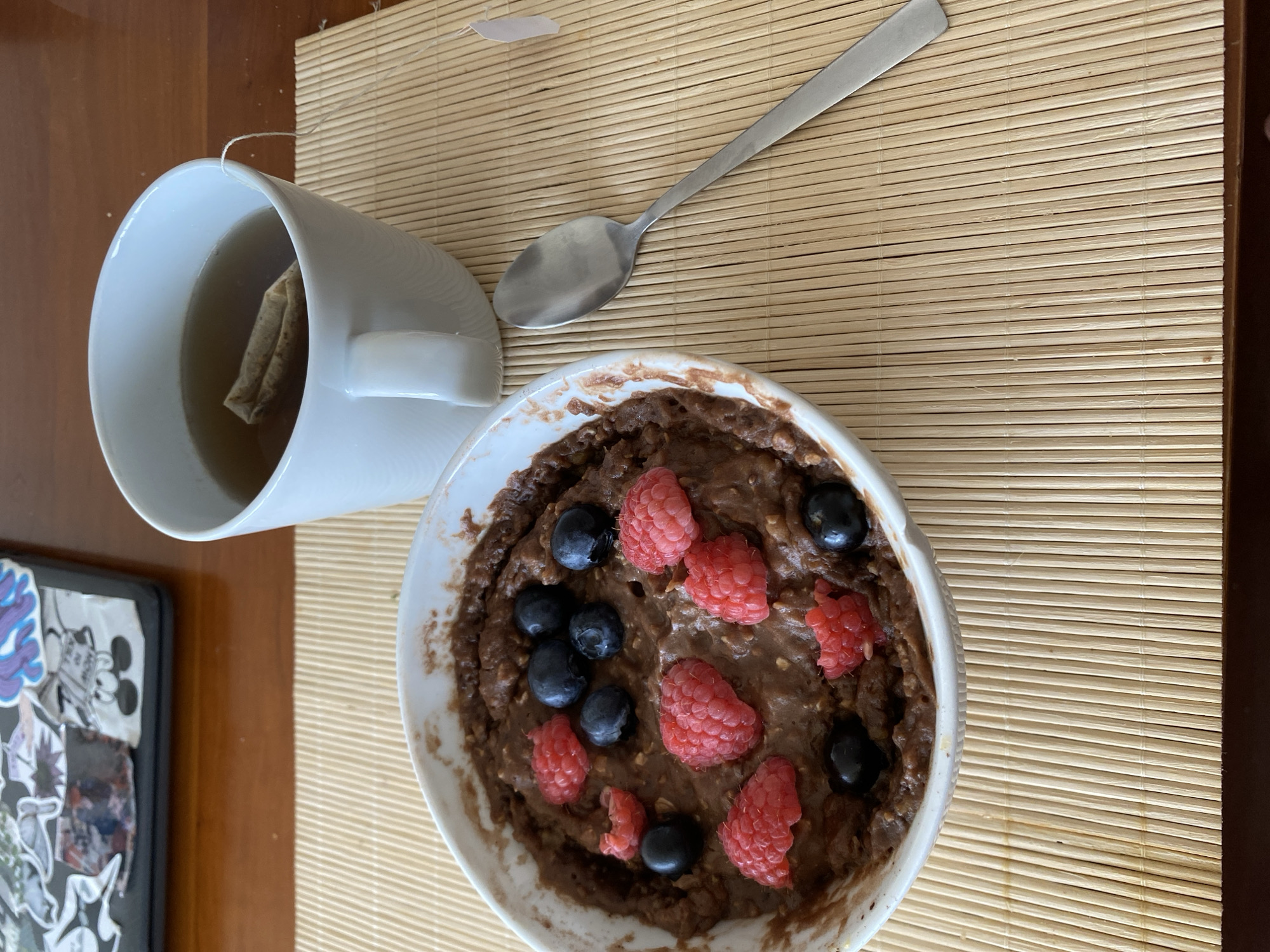 A top-down shot captures a breakfast scene on a wooden table covered by a woven bamboo placemat. In the foreground, a white bowl is filled with chocolate oatmeal, garnished with fresh raspberries and blueberries. To the left of the bowl, a white mug contains tea with a tea bag submerged. A silver spoon rests on the placemat, positioned between the mug and the bowl. Partially visible on the lower left is a black laptop adorned with various stickers. The lighting suggests it's daytime, and the overall mood is relaxed and domestic. No discernible text is visible in the image.