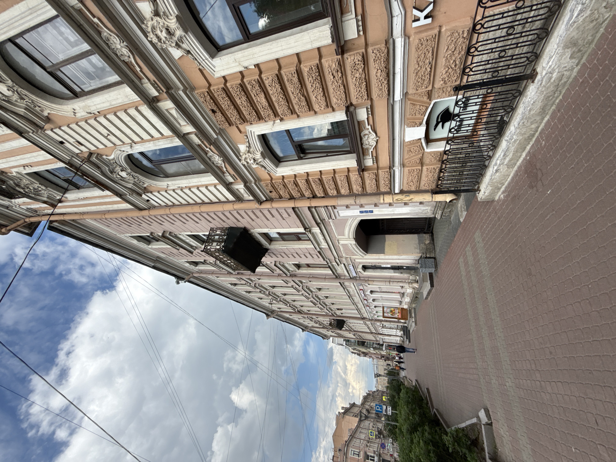 The image depicts a street scene with a focus on an ornate building facade on the right and a paved sidewalk extending into the distance on the left. The building, constructed with a pale pinkish-beige base and extensive white architectural details, features intricate moldings, decorative cornices, and textured wall sections. Several windows, both arched and rectangular, are visible, some reflecting the cloudy sky.
A black wrought-iron balcony railing is present on an upper floor, near a white sign with a black silhouette of a bird. Beneath the bird, the text "МАЛЕНЬКИЙ БАР" (Small Bar) is partially visible. A large white letter "K" is also affixed to the building's facade. Further down the street, an arched entryway can be seen.
The sidewalk is paved with reddish-brown blocks in a distinct pattern. A few pedestrians are visible walking in the distance on the sidewalk. Green trees or bushes line the edge of the street further down. Overhead, numerous thin wires crisscross against a bright blue sky dotted with scattered white clouds. The scene suggests a daytime view of a historic city street, likely on a partially cloudy day.