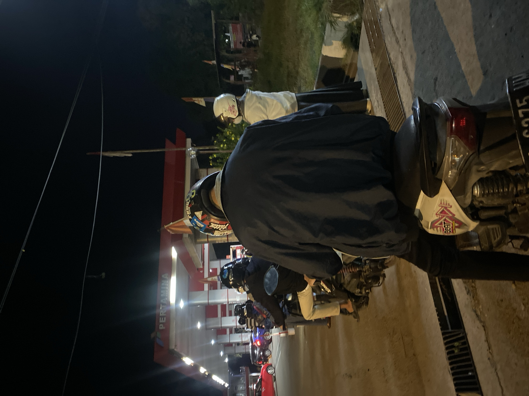 The image depicts a night scene at a Pertamina gas station in Gambut Barat, Indonesia. Several motorcycles are lined up, with people on them, presumably waiting to refuel. The gas station canopy is illuminated, and the Pertamina logo is visible. The foreground is dominated by the back of a motorcycle and its rider, who is wearing a dark jacket and helmet. In the background, other vehicles, including cars and motorcycles, are present. A woman wearing a white jacket and a long dark skirt, with a white helmet adorned with a floral pattern, stands near a motorcycle. The overall atmosphere is that of a busy, everyday scene at a local gas station at night.