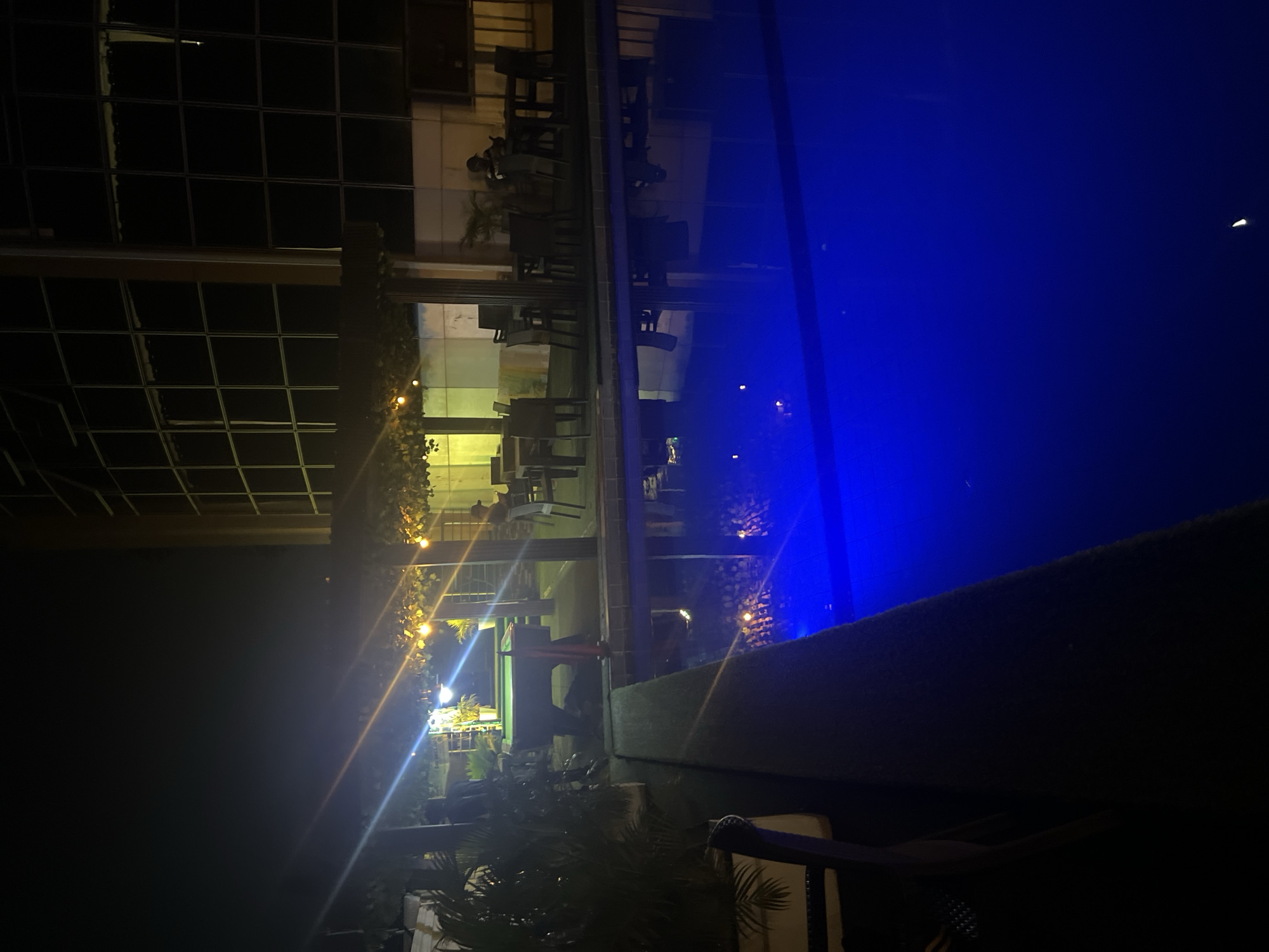 The image displays a nighttime scene of an outdoor leisure area in Abuja, Nigeria, characterized by vibrant artificial lighting. The composition is horizontally oriented after mental correction.
On the right side, a prominent, large swimming pool or water feature glows with an intense, uniform blue illumination, creating a striking contrast against the dark surroundings. A low, dark barrier or wall runs across the foreground, separating the viewer's perspective from the water body.
To the left of the pool, an outdoor dining or lounge area is visible. This space features multiple dark tables and chairs, arranged as if for patrons. It appears to be an extension of an indoor structure, visible through large glass panels or windows that reflect some of the ambient light. Various warm-toned artificial lights, including what appear to be string lights, illuminate this area, creating notable lens flares and starburst effects within the frame.
Scattered greenery, consisting of potted plants or bushes, is present around the dining area and along the architectural elements. The structures on the far left exhibit a grid-like pattern, likely from window panes or a facade. The time of day is clearly night, with no natural light evident. The atmosphere conveyed is peaceful and inviting, typical of an evening at an upscale establishment. No clearly identifiable people or specific activities are depicted, and there is no visible text in the image.