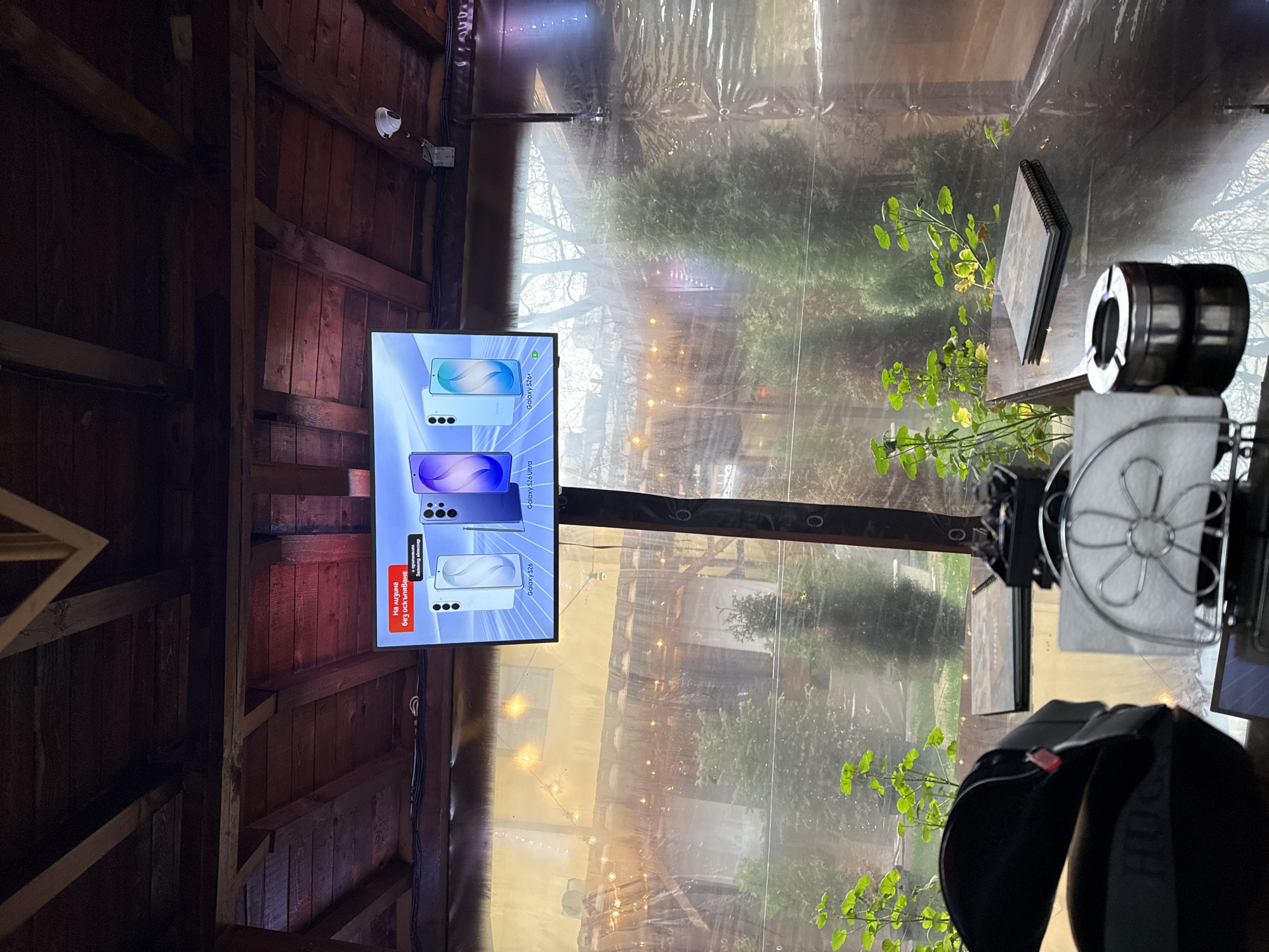 The image shows the interior of an enclosed space, likely an outdoor seating area of a restaurant or bar, with a wooden structure visible on the left. A flat-screen TV is mounted on the wood, displaying what appears to be advertisements for Samsung Galaxy S26 smartphones.
Through a semi-transparent barrier, one can see an outdoor area with trees and lights, suggesting a patio or garden. The lighting indicates it could be daytime or early evening.
Objects such as a menu holder and a fan are also in the scene, providing context to the location being a dining or recreational space.
The OCR text within the advertisement displays the phrases “На лизинг без оскъпяване” which translates to "leasing without appreciation," and the models "Galaxy S26," "Galaxy S26 Ultra," and "Galaxy S26+".
There are no people visible in the image. Given the language used in the advertisement, it's highly probable that this scene is located in Bulgaria, with the most likely city being Sofia.