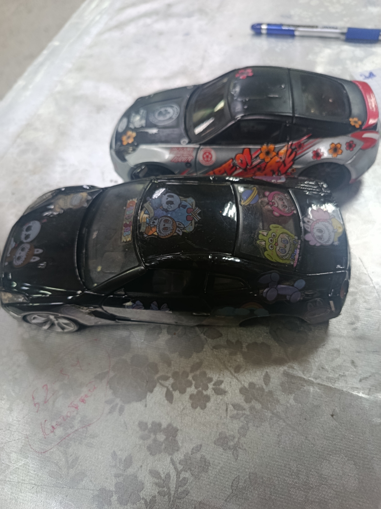 The image shows two toy cars on a patterned tablecloth. The car in the foreground is black and decorated with stickers of cartoon characters. The car in the background is silver and black, with graphics of flowers and graffiti. A blue pen is visible in the upper right corner. The setting is not identifiable as Sertolovo, Russia.