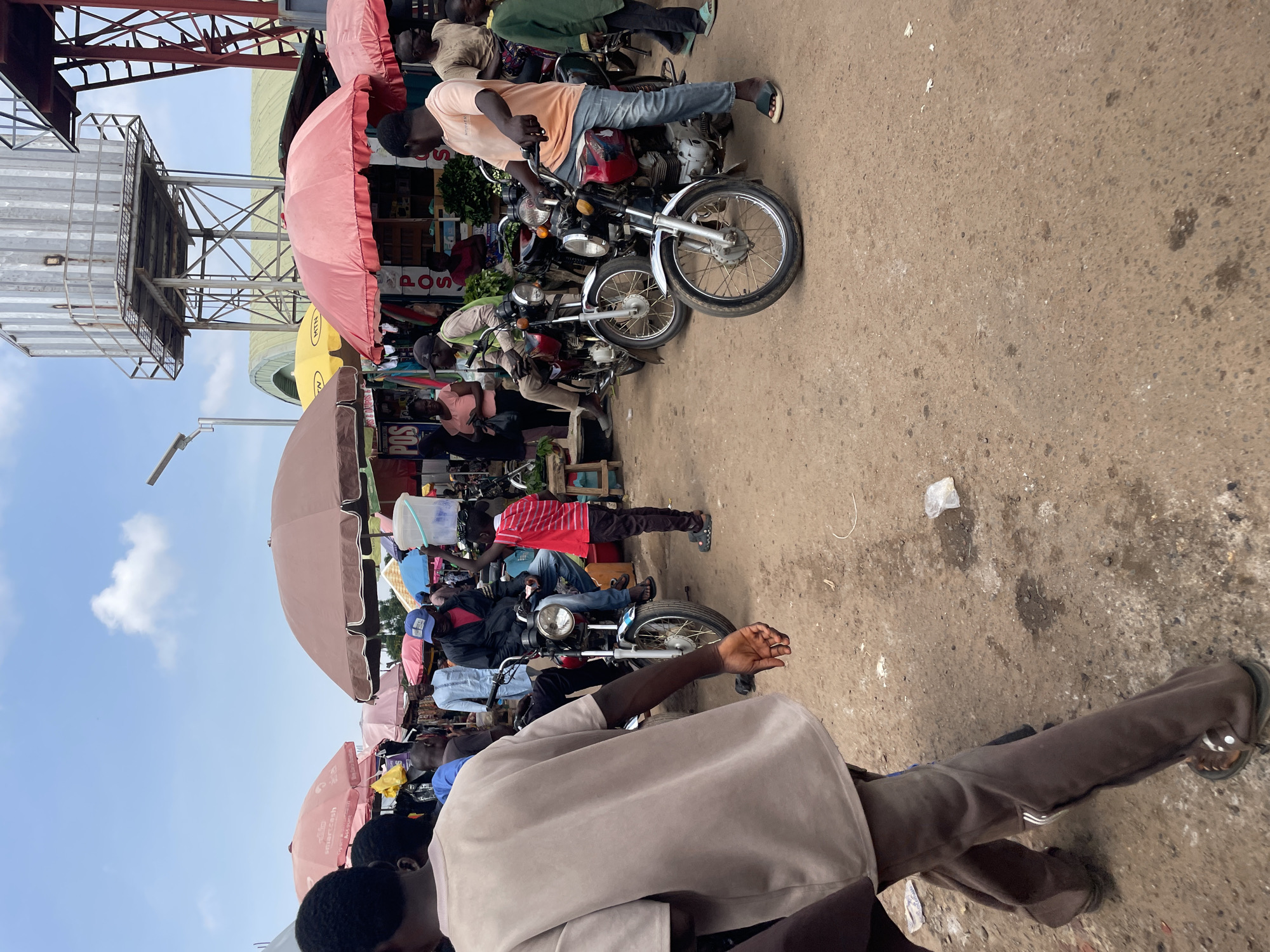 The image depicts a bustling street scene in Akwanga, Nigeria, likely during the day under a bright sky with scattered clouds. The ground is a mix of dirt and concrete, showing signs of wear and tear, with some scattered debris.
Several people are present, primarily young men, engaged in various activities related to what appears to be a marketplace or transportation hub. Many are associated with motorcycles, which are lined up in rows. Some individuals are seated on the motorcycles, while others are standing or walking nearby. One person is seen holding a large white bucket. The people are dressed in casual attire, reflecting a warm climate. Their expressions are not clearly discernible, but the overall atmosphere suggests a busy, everyday scene.
A prominent feature of the scene is the presence of numerous large, colorful umbrellas, primarily pink and red, providing shade. These umbrellas are likely part of market stalls. In the background, there are structures that could be market stalls or buildings, including metal scaffolding and what looks like a part of a utility pole or antenna.
There are some visible text elements: "POS" is visible on a sign, likely indicating Point of Sale. The "MTN" logo is also visible on a yellow umbrella, indicating a telecommunications company.
The orientation of the photo is rotated, with the subjects and scene appearing sideways. The activity seems to be centered around the motorcycles, suggesting they might be used for transportation, possibly as motorcycle taxis, or for transport of goods. The setting appears to be an open-air market or a busy street intersection where people gather.