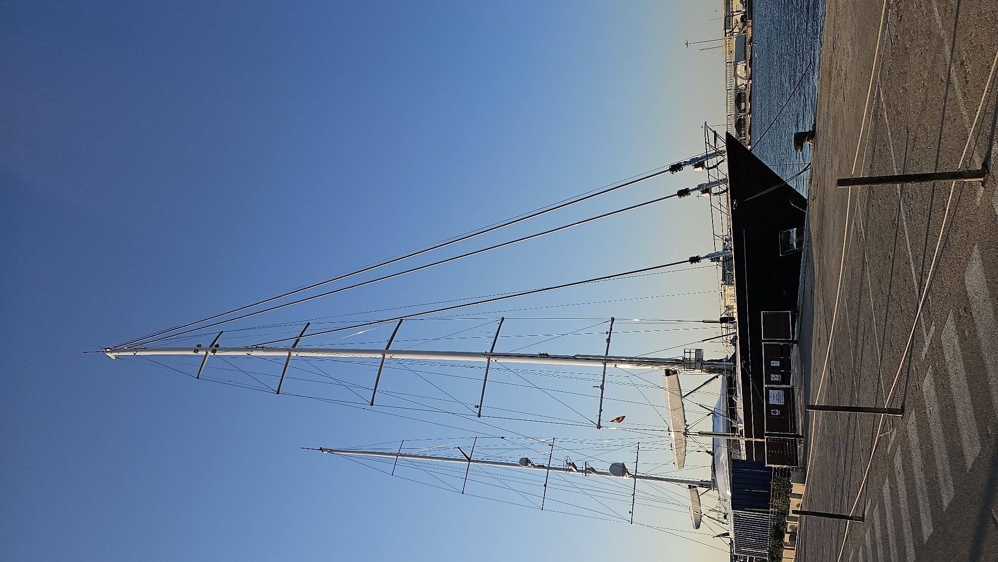 The image shows a large black sailboat docked at a pier in the city of Dénia, Spain. The boat has multiple tall masts and rigging against a clear blue sky. The pier is paved with a crosswalk painted in white stripes in the foreground, with bollards and ropes marking off sections of the docking area. In the background, a bridge can be seen spanning across the water, and industrial buildings line the distant shore. The water is calm, reflecting the clear sky. No people are visible in the image.