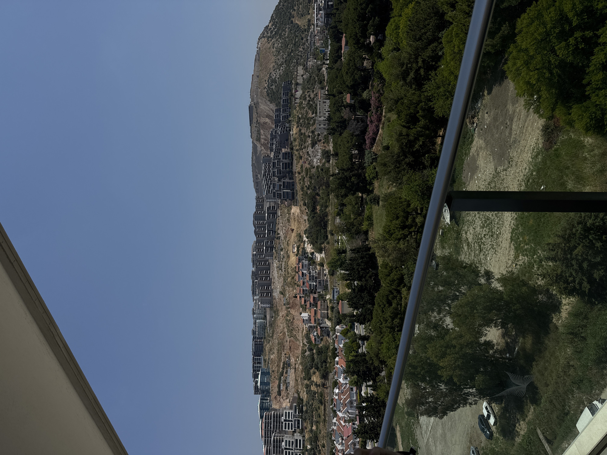 The image presents a high-angle panoramic view, likely from a balcony or elevated structure, looking out over a landscape in Kuşadası, Turkey, under a clear, bright blue sky.
In the immediate foreground, a dark grey or black metal railing with a transparent glass panel extends horizontally across the lower portion of the frame. A faint, reflective emblem, possibly resembling wings or a bird, is visible on the glass panel to the left. The top edge of a light-colored building structure is partially visible in the upper left corner, indicating the vantage point.
Below the railing, the immediate ground consists of varied terrain. On the left, a dirt or unpaved area functions as a parking lot, where a black sedan and a white sedan are parked. Sparse, dry vegetation and patches of green trees are scattered across this section. Towards the bottom edge, a small portion of a white vehicle is visible.
Moving into the mid-distance, a residential area is visible, characterized by clusters of houses with light-colored facades and reddish-orange tiled roofs, interspersed with numerous green trees and shrubs. Roads are discernible winding through these settlements.
The background is dominated by a series of prominent hills. These hills are extensively developed with a high density of modern, multi-story buildings, predominantly featuring grey and white facades. Many structures appear to be newly constructed or actively under development, indicated by visible construction sites and significant areas of exposed reddish-brown earth from excavation and grading. Patches of sparse, dry vegetation and trees are also present on the less developed slopes.
The sky is uniformly clear and cloudless, indicating sunny weather. No individuals are visible in the scene. The overall impression is one of urban expansion into a hilly, natural landscape.