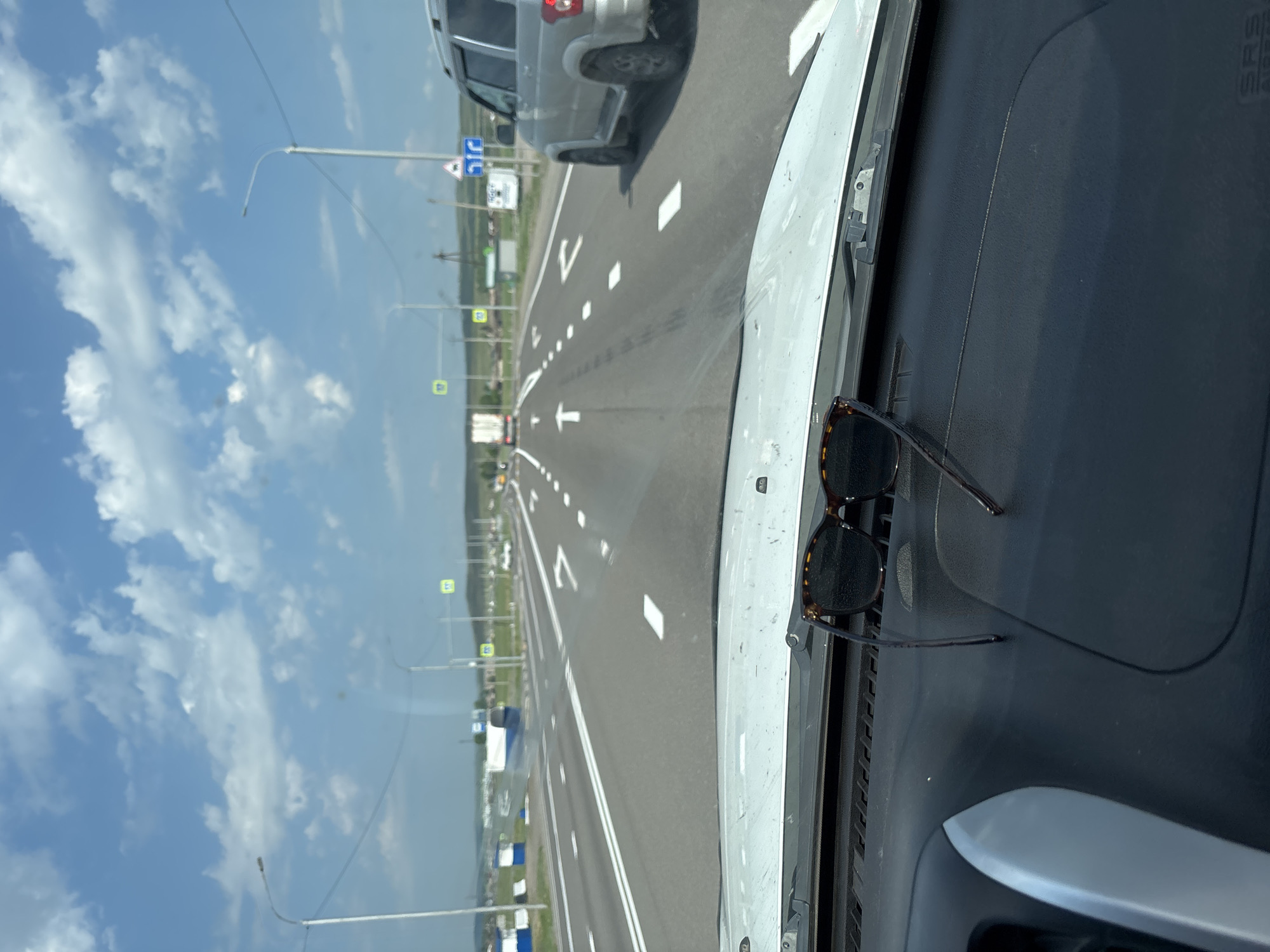 The image captures a view from inside a vehicle, looking forward through the windshield. Sunglasses with tortoiseshell frames are placed on the dashboard in the foreground. The road ahead is a multi-lane highway with white lane markings. Several vehicles, including a truck and an SUV, are visible on the road. The environment appears to be rural, with fields and scattered buildings visible on either side of the highway. The sky is blue with scattered white clouds. The location is identified as Troitskoye, Russia.