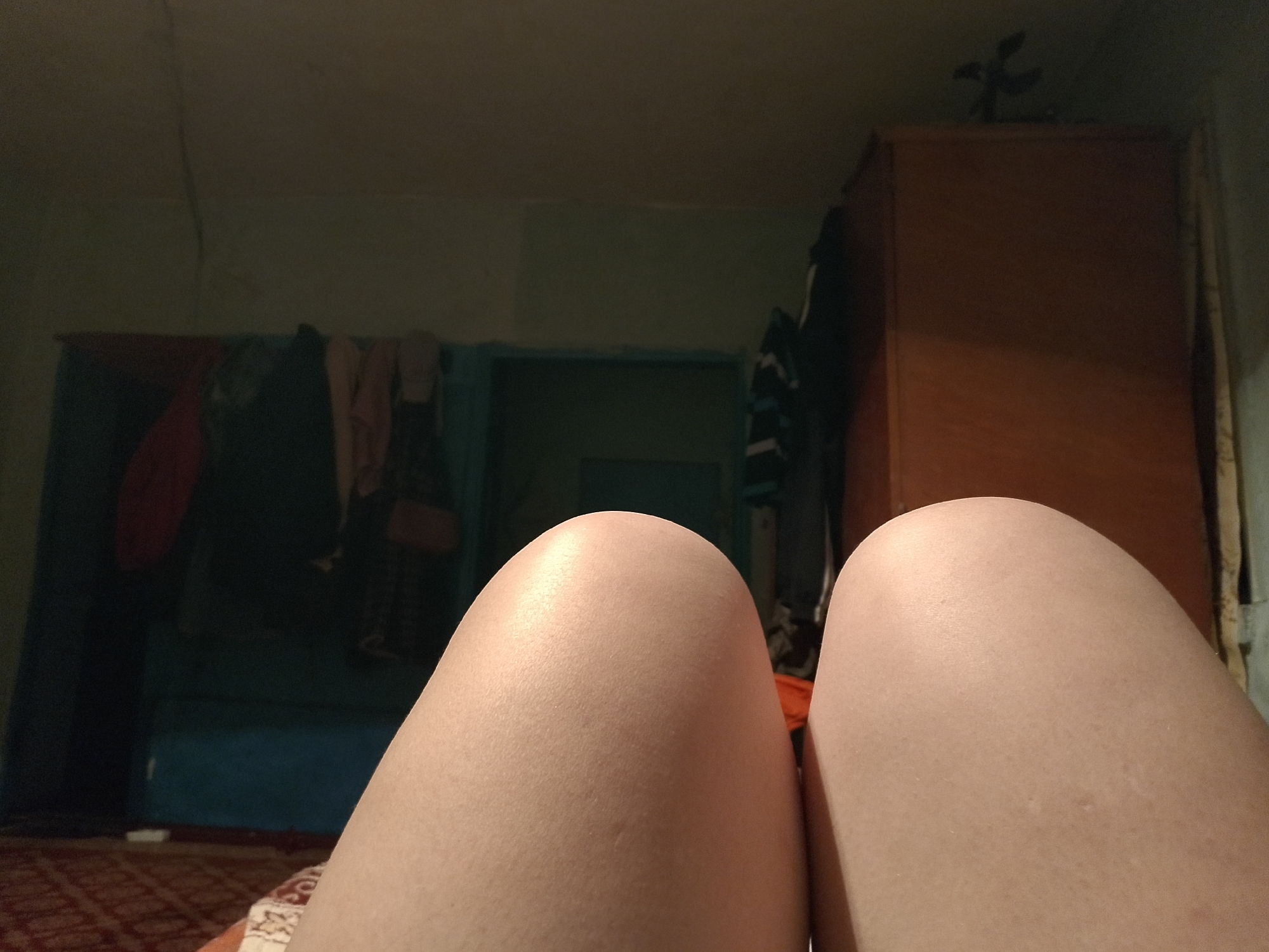 The image depicts an indoor scene from a first-person perspective, looking out from a seated or reclined position in a room in the city of Unknown, Unknown. The lower half of the frame is dominated by the upper thighs of a person, appearing smooth and light-skinned, suggesting a relaxed posture. The background reveals a dimly lit room with muted colors. To the left, a wall painted in a faded light green or beige hue features a dark blue/green partition or structure, behind which various garments, including a red item and dark clothing, are visible hanging. A section of a red patterned rug is partially visible on the floor in this area. To the right, a tall, brown wooden wardrobe or cabinet stands against the wall. A dark, indistinct object, possibly a decorative item or plant, is positioned on top of the cabinet. A light-colored fabric or curtain runs vertically along the right edge of the frame. The overall lighting is low, creating a subdued atmosphere within the room.