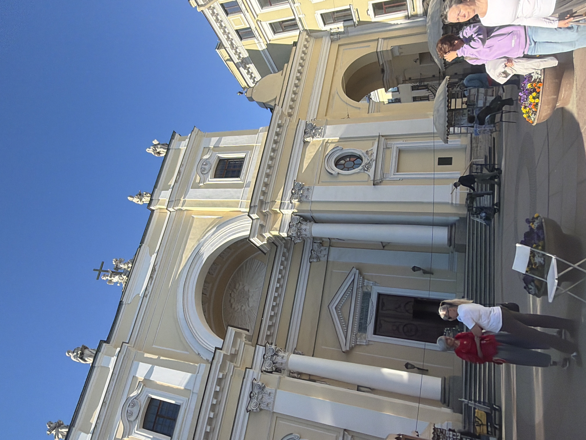 This is a photograph of the exterior of a building, likely a church or historical building, in Saint Petersburg, Russia. The building is predominantly light yellow with white architectural details. It features a large arched entrance, ornate columns, and decorative carvings. Several statues are visible on the roofline and facade, including one with a cross. The sky is a clear, bright blue, suggesting a sunny day.
In the foreground and midground, several people are visible. Two women are standing near a large planter filled with colorful flowers, one wearing a white top and the other a purple hoodie. Further back, there are other individuals and what appear to be market stalls or outdoor seating areas, some with umbrellas. One person in the foreground is wearing a red shirt and is near another person in a white top. The overall atmosphere seems relaxed, with people enjoying the outdoors. The time of day appears to be daytime, given the bright sunlight casting shadows.