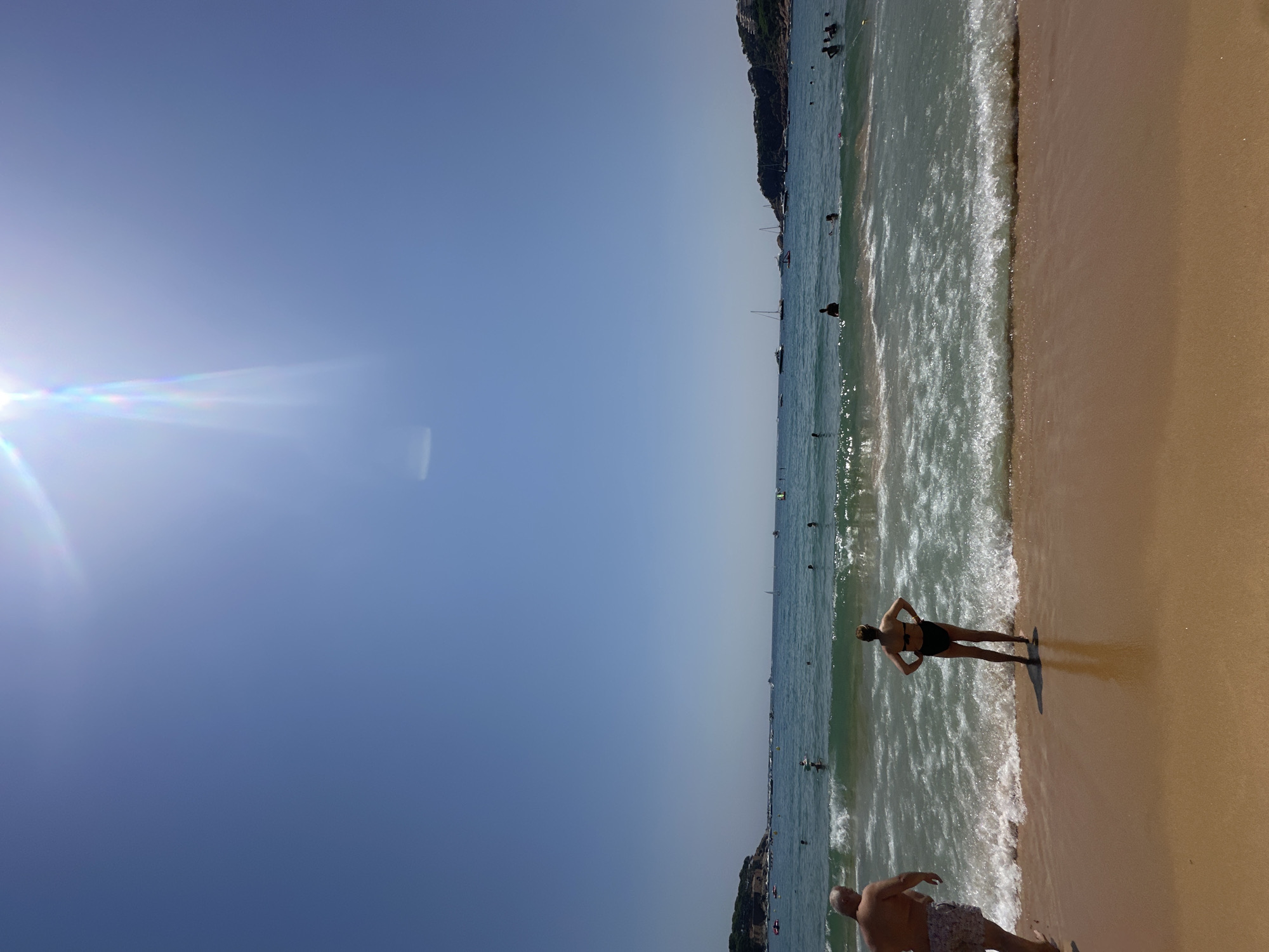 The image depicts a sunny day at a beach in Sant Feliu de Guíxols, Spain. In the foreground, a sandy beach meets the ocean, with gentle waves lapping at the shore. Two individuals are visible: a man on the left facing away from the camera and a woman in a black bikini standing with her hands on her hips, facing the water. The sea is populated with numerous people swimming and boating, including several sailboats and yachts anchored in the distance. Lush green hills with some buildings are visible on the coastline in the background, framing the bay. The sky is clear and blue, with the sun positioned high and slightly to the right, casting bright lens flares and a slight haze. The overall scene is one of a lively and picturesque coastal resort.