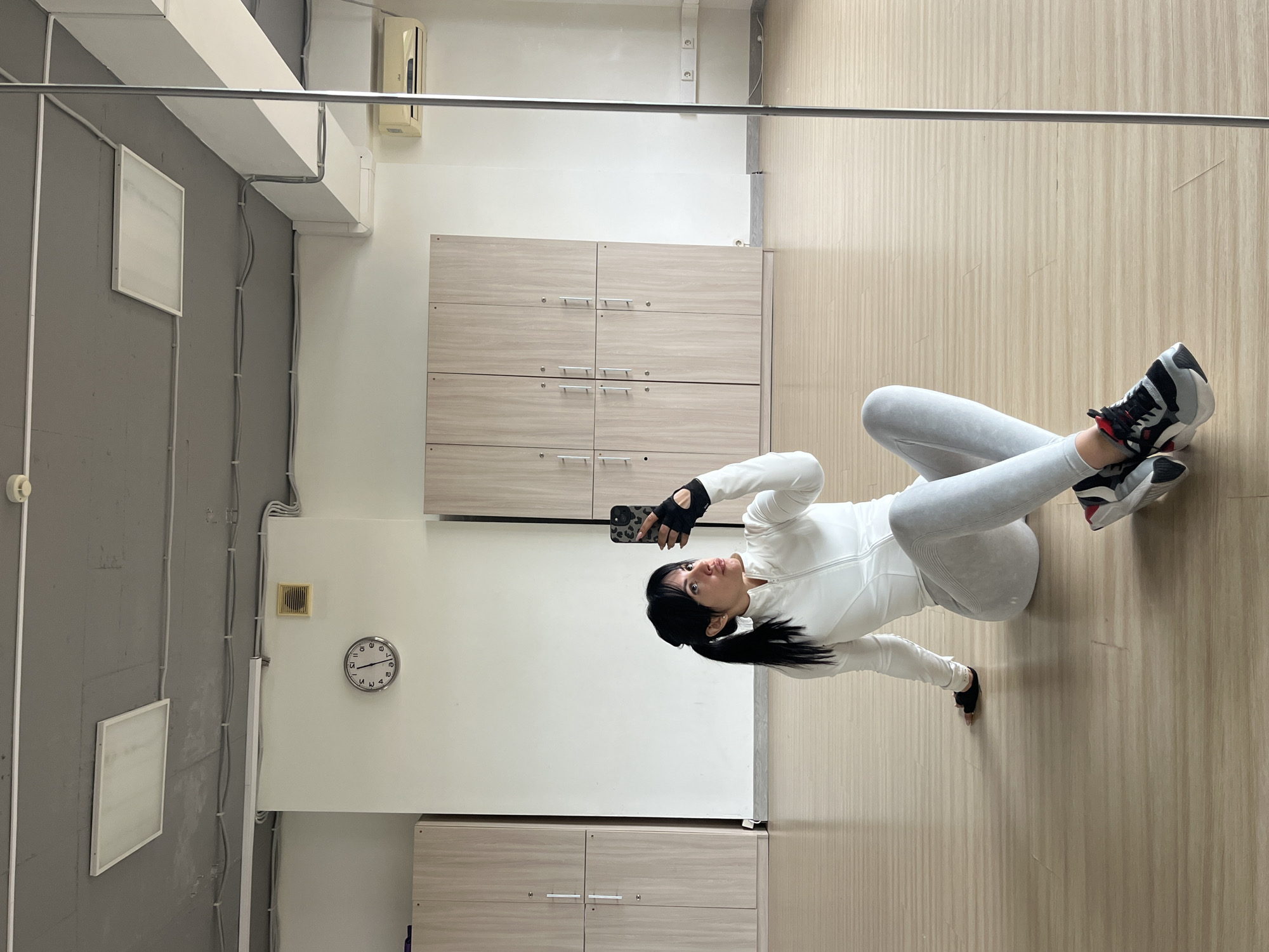 A person in a white zip-up jacket and gray leggings is taking a selfie in a room. The room has a light wood floor, white walls, and light wood cabinets. A clock on the wall reads approximately 1:55. There are also visible pipes and electrical conduits along the wall and ceiling. The location cannot be determined from the image.