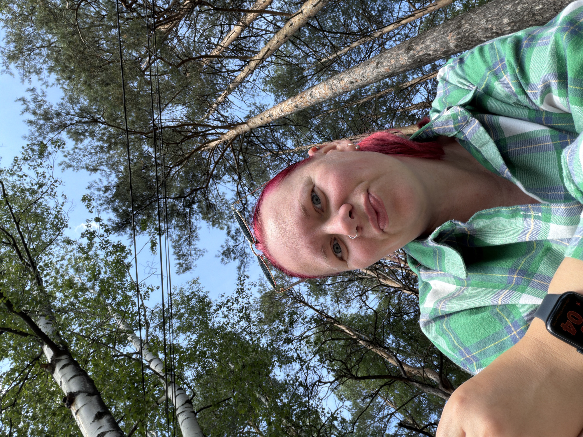 A low-angle selfie captures a light-skinned individual with short, bright pink hair, light eyes, a nose ring in the left nostril, and sunglasses perched on their head. They are wearing a green, white, and blue plaid shirt, with a dark smartwatch displaying "04" on their left wrist. The individual looks directly at the camera with a subtle smirk. The background features a clear blue sky, framed by tall trees, including birches with white bark and green leaves, and coniferous pine trees with needle-like foliage. Multiple dark power lines traverse horizontally across the upper middle section of the sky. The scene is situated in the city of Chaikovsky, Russia.