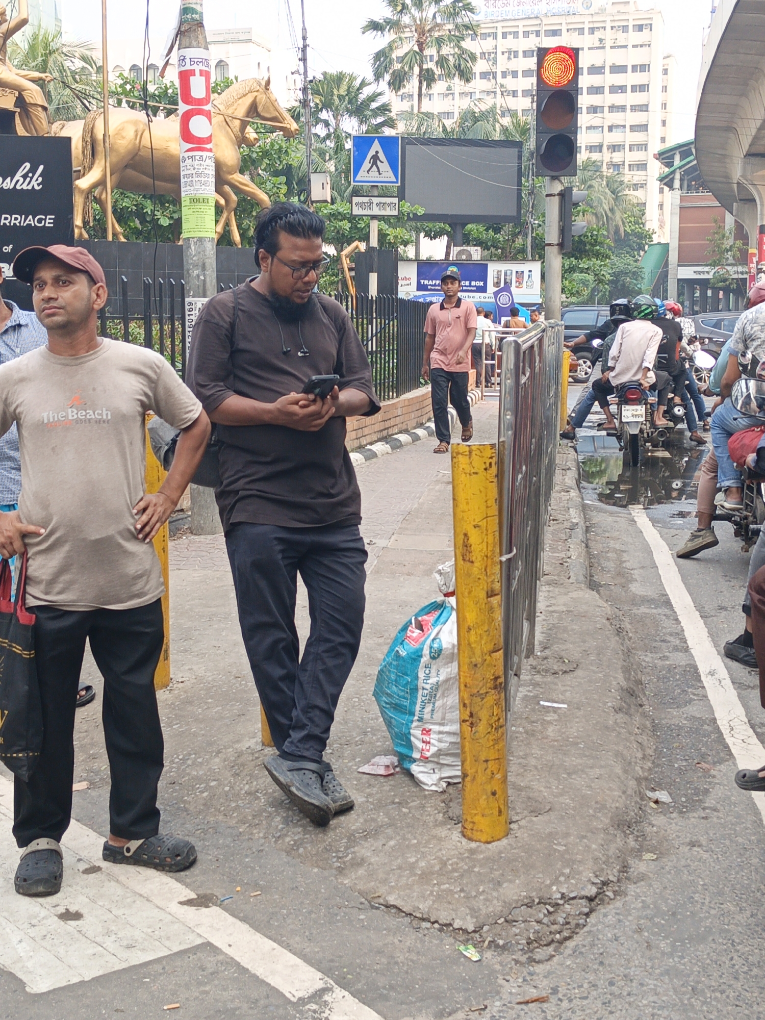 The image captures a busy urban scene in Unknown, Bangladesh. Two men stand on the sidewalk, one wearing a red cap and a light brown t-shirt with the words "The Beach GOES HERE" and the other wearing glasses, a black t-shirt, and holding a smartphone. A white and blue bag labeled "TEER MINIKET RICE 25 kg PREMIUM QUALITY" sits next to a yellow pole.
Behind the men, a traffic signal shows a red light, and a pedestrian crossing sign is visible. Further back, a golden horse statue sits atop a structure behind a black fence. Motorcycles and cars are stopped in the road, partially obscured by a puddle. A man in a pink shirt walks on the sidewalk in the background, and a multi-story building is visible in the distance. Signs with Bengali script are present on poles and buildings.