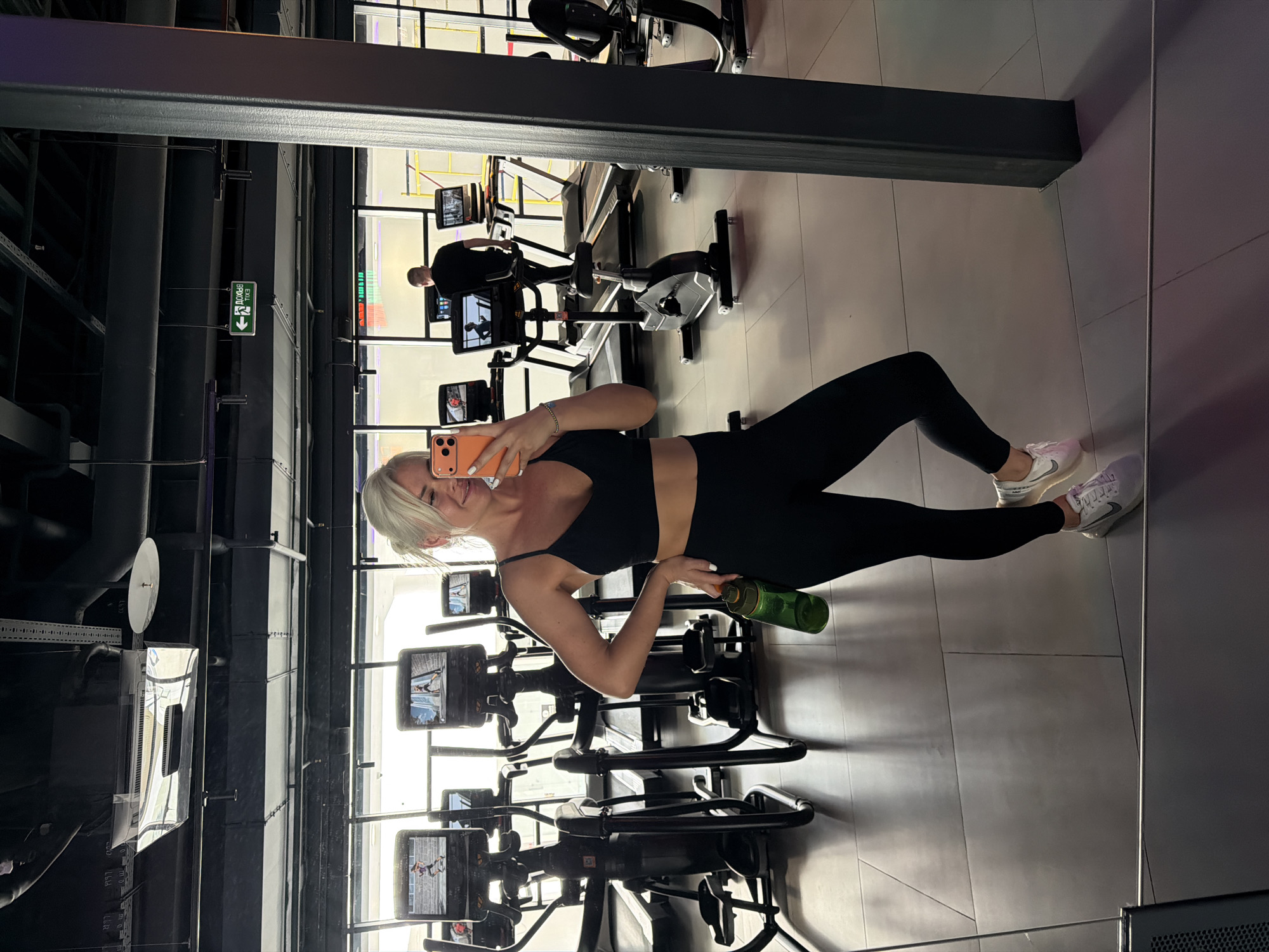 A woman is standing in a gym, taking a selfie in a mirror. She is wearing a black sports bra and leggings, and white sneakers. She is holding a green water bottle. In the background, there are several exercise machines, including stationary bikes and a treadmill. There is a man on one of the exercise machines. The location appears to be a modern gym with black and gray accents. There is a sign on the wall. The location is in the city of Unknown, Unknown.
