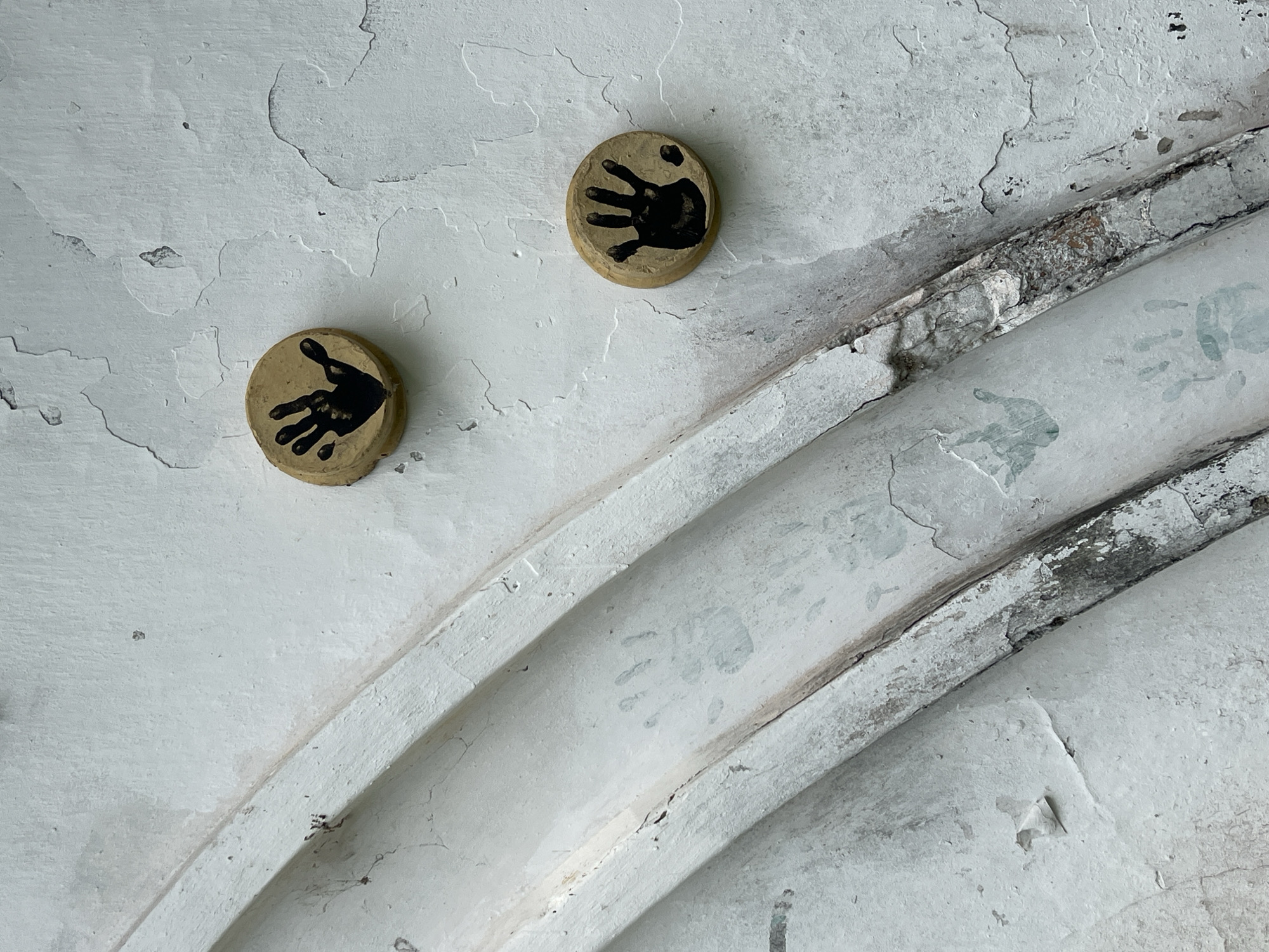 A close-up view reveals a textured, light-colored wall, likely painted white or off-white, showing considerable wear with peeling paint and visible cracks. Two circular, light brown discs are affixed to the wall in the upper-left quadrant. Each disc features a distinct black handprint; the handprint on the upper-right disc is slightly less defined than the one on the lower-left.
The wall's surface is uneven, suggesting plaster or concrete. Diagonally traversing the right side of the frame are two parallel, raised, curved structures, possibly pipes or architectural moldings. These structures also show signs of extensive wear, with chipped paint and exposed darker material underneath. Faint, faded handprints are subtly visible on the surface of the lower-right curved structure, integrating with the distressed texture of the wall. The scene evokes a sense of age and past human interaction, with the handprints serving as a poignant detail. There is no visible text.