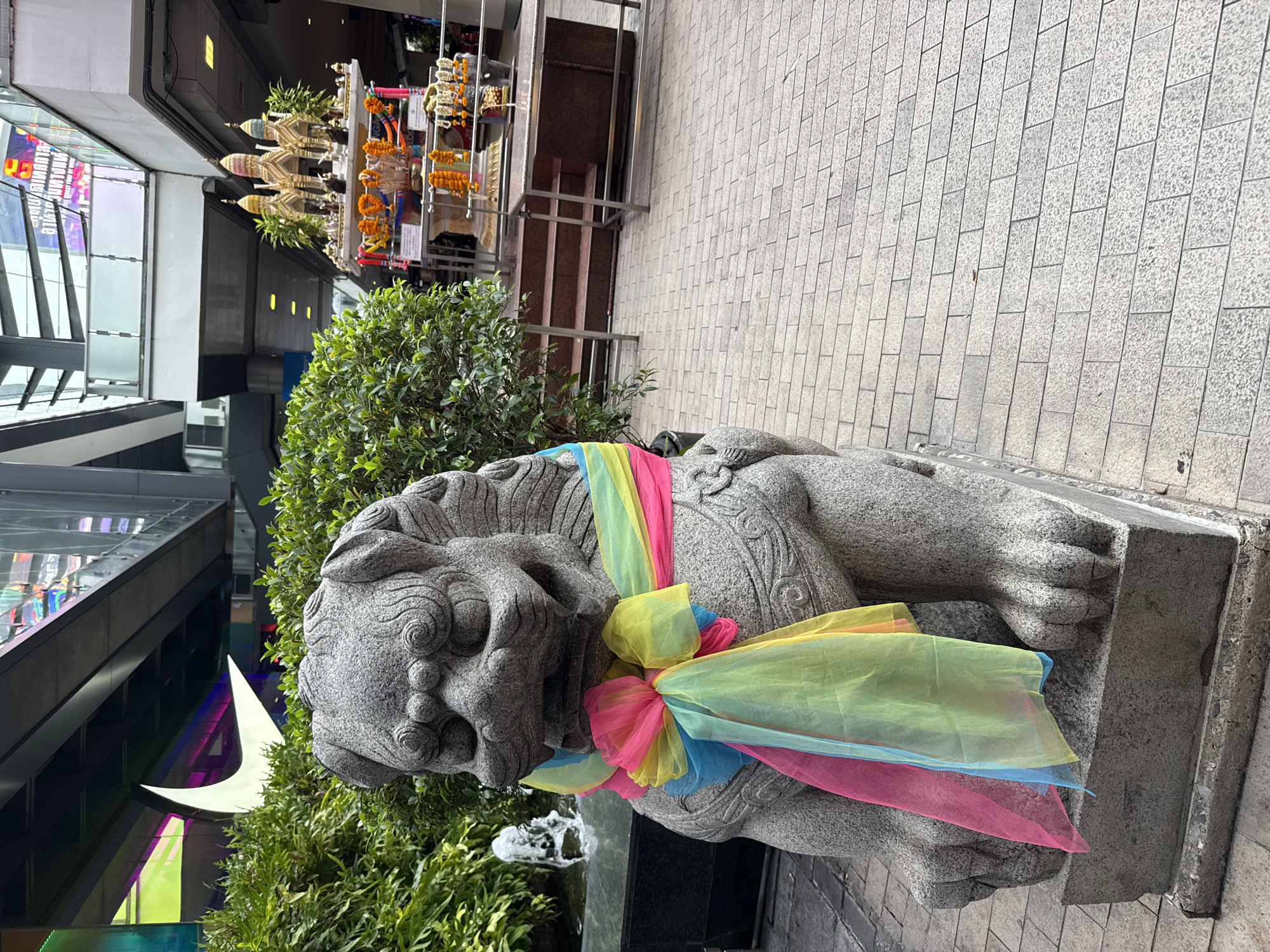 A stone statue of a lion, adorned with colorful sheer fabrics, is positioned on a concrete base. Lush green foliage surrounds the base. In the background, a multi-tiered structure with golden ornaments is visible, possibly a shrine. The setting is outdoors, with grey pavement and a partial view of modern architecture featuring glass and steel. This appears to be a commercial or public space in Bangkok, Thailand, where such shrines and statues are common.