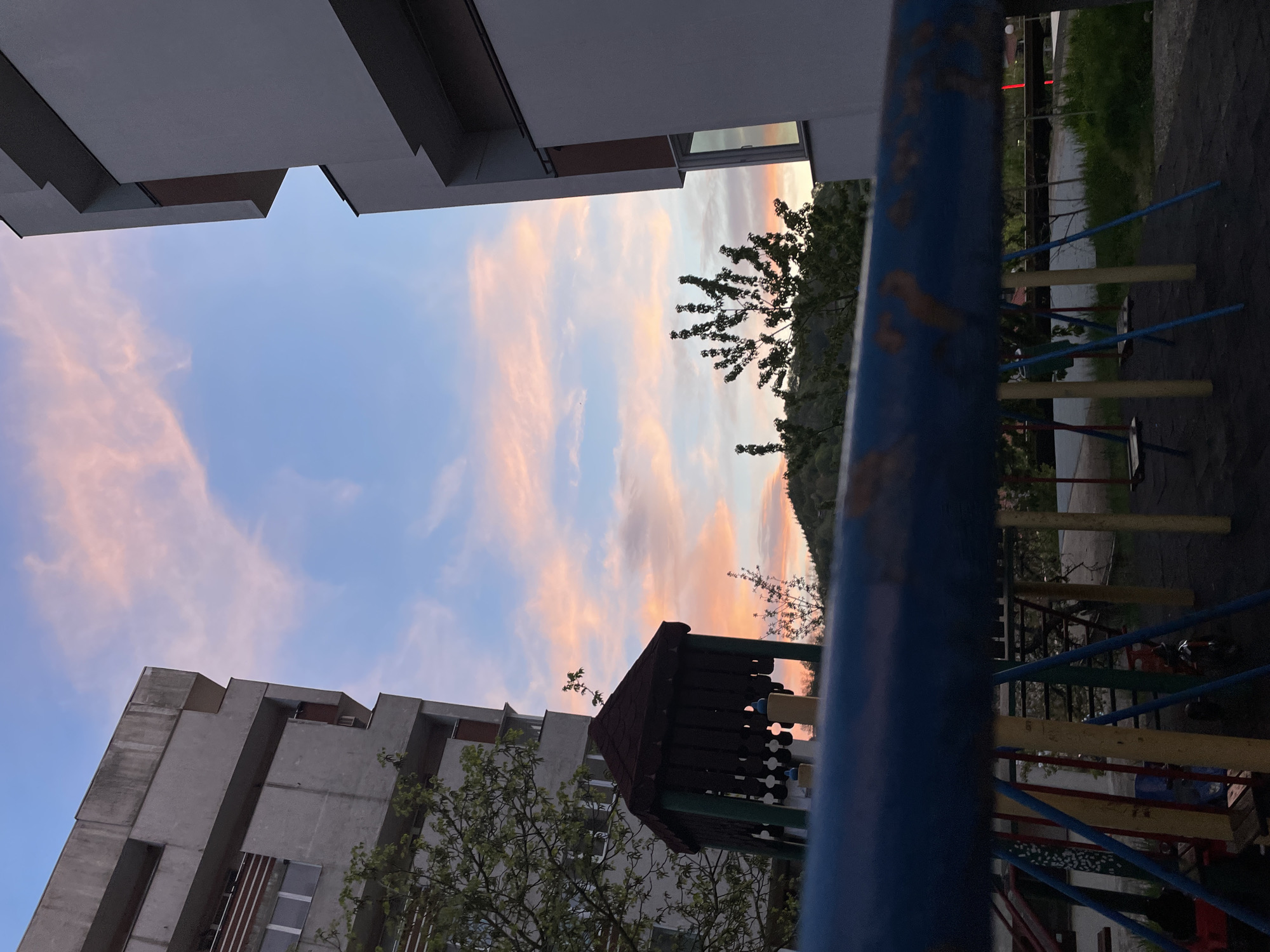 The image shows a playground in Zlatograd, Bulgaria, at dusk. The sky is a mix of blue and pink hues. The playground includes a small playhouse and various climbing structures constructed from blue, yellow, and red painted poles. A large blue pole is in the foreground, partially obscuring the view. In the background, there are two large concrete buildings, framing the sky. A tree with sparse leaves is also visible. Beyond the playground, some green foliage suggests a park or grassy area. There are no people visible in the image. The setting appears to be a residential area.