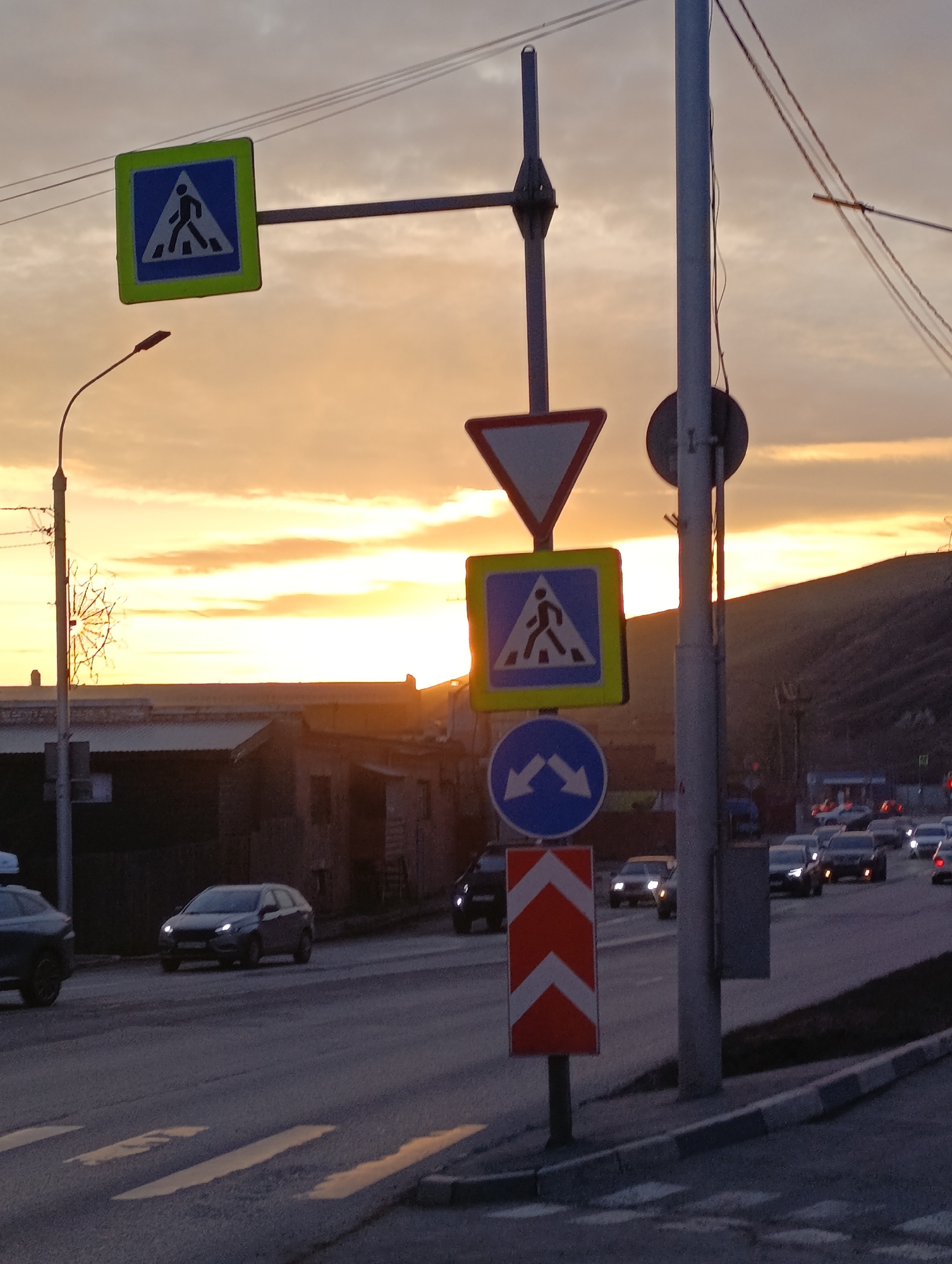 The image shows a street scene at sunset in Krasnoyarsk, Russia. A series of road signs are prominent in the foreground, including pedestrian crossing signs and a directional arrow sign. A street is visible with several cars and crossing. The buildings and terrain in the background are silhouetted against the bright sunset sky. There are no visible people.