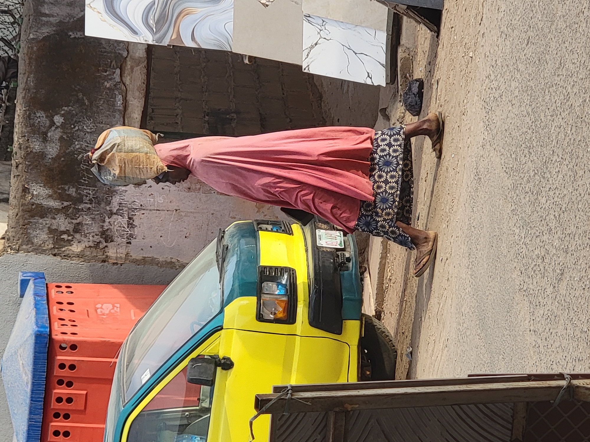 In Maiburiji, Nigeria, an older woman wearing a head covering, a pink long-sleeved garment, and a patterned skirt stands next to a yellow and blue truck. The woman faces left and appears to be walking. A stack of orange crates is to the left of the truck's front. Tiles are leaning against a wall behind the woman. The ground is paved.