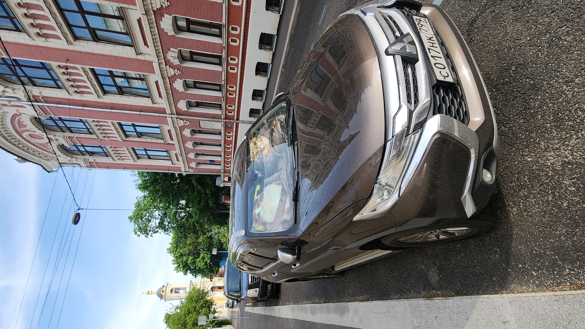 The image depicts a brown Mitsubishi Outlander SUV parked on a street in Moscow, Russia. The license plate reads "C017HK 799." Inside the car, two fluffy cats are visible in a carrier on the front passenger seat. The vehicle is parked next to a curb and another car is seen behind it. The background includes a red brick building with white accents and numerous windows, as well as trees, a church with a golden dome and cross, a street lamp, and a blue sky. The pavement is asphalt.