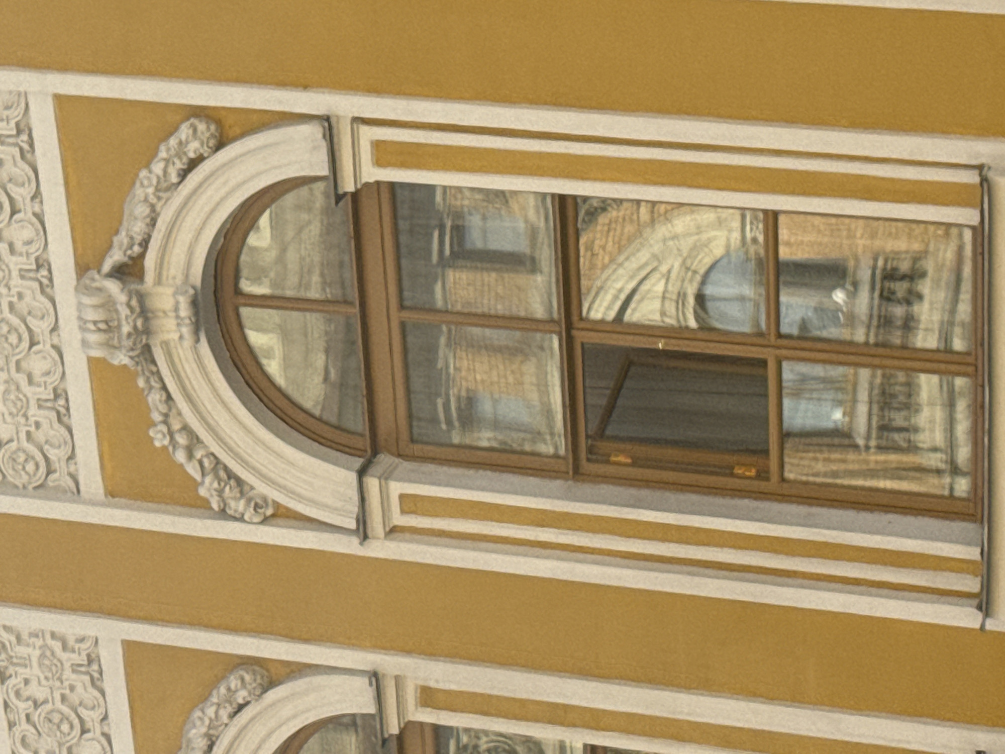 The image shows a close-up of an arched window on a building with yellow walls and white decorative trim. The window is framed by dark brown wood and divided into multiple panes by the same dark brown material. The glass reflects the facade of another building, featuring brickwork and arched windows. The reflections also show ornate architectural details. The building in the foreground displays decorative stucco elements above the arch of the window, consisting of floral motifs and a central crest. The overall architectural style suggests a historical building, possibly in the city of Saint Petersburg, Russia, known for its ornate classical and baroque architecture.