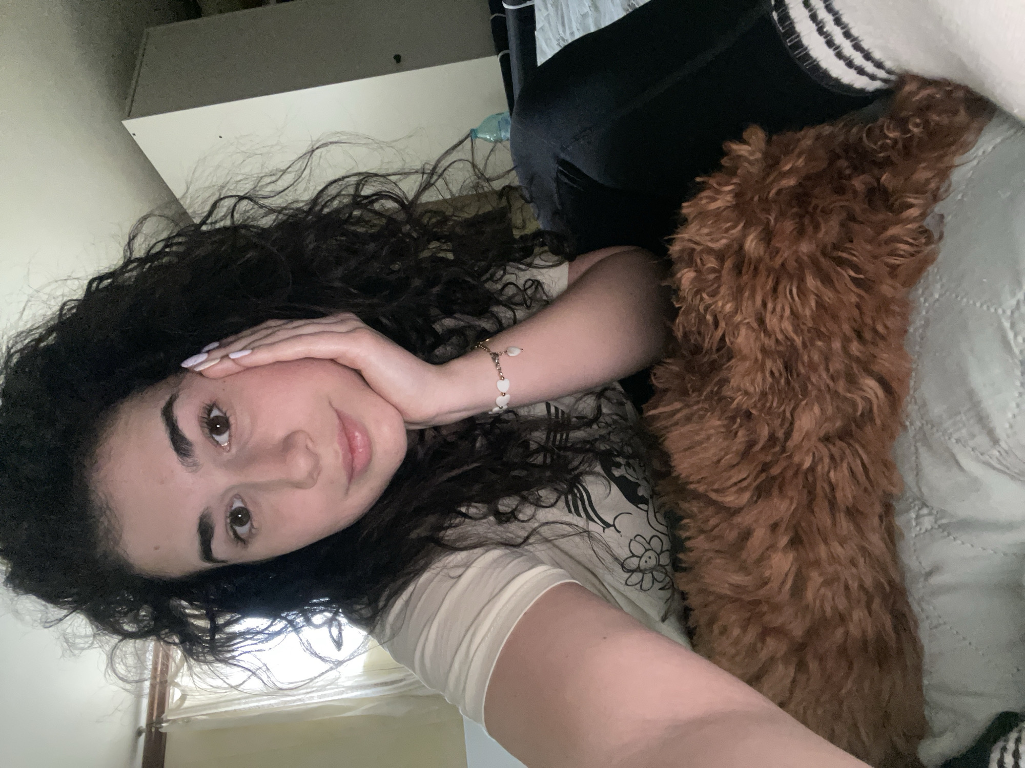 The image, taken in Sofia, Bulgaria, displays a candid indoor scene featuring a young woman and a reddish-brown dog. The woman, with long, dark, curly hair, looks directly at the camera with a gentle expression and a slight smile. Her head is tilted, and her left hand, adorned with pale-colored nails and a white beaded bracelet, rests softly against her cheek. She is wearing a light-colored t-shirt with a floral graphic that includes a cartoon-like smiling flower.
Alongside her, a dog with dense, curly, reddish-brown fur is partially visible on the right side of the frame, suggesting it is lying close to her, possibly nestled on a bed or sofa. The dog's breed appears to be a poodle or similar curly-haired type.
The background shows a simple interior with a light-colored wall, a light-toned textured blanket or quilt, and hints of a window with blinds or curtains. The lighting is soft and natural, indicating daytime. The overall atmosphere conveyed is one of relaxation, comfort, and affection between the two subjects.