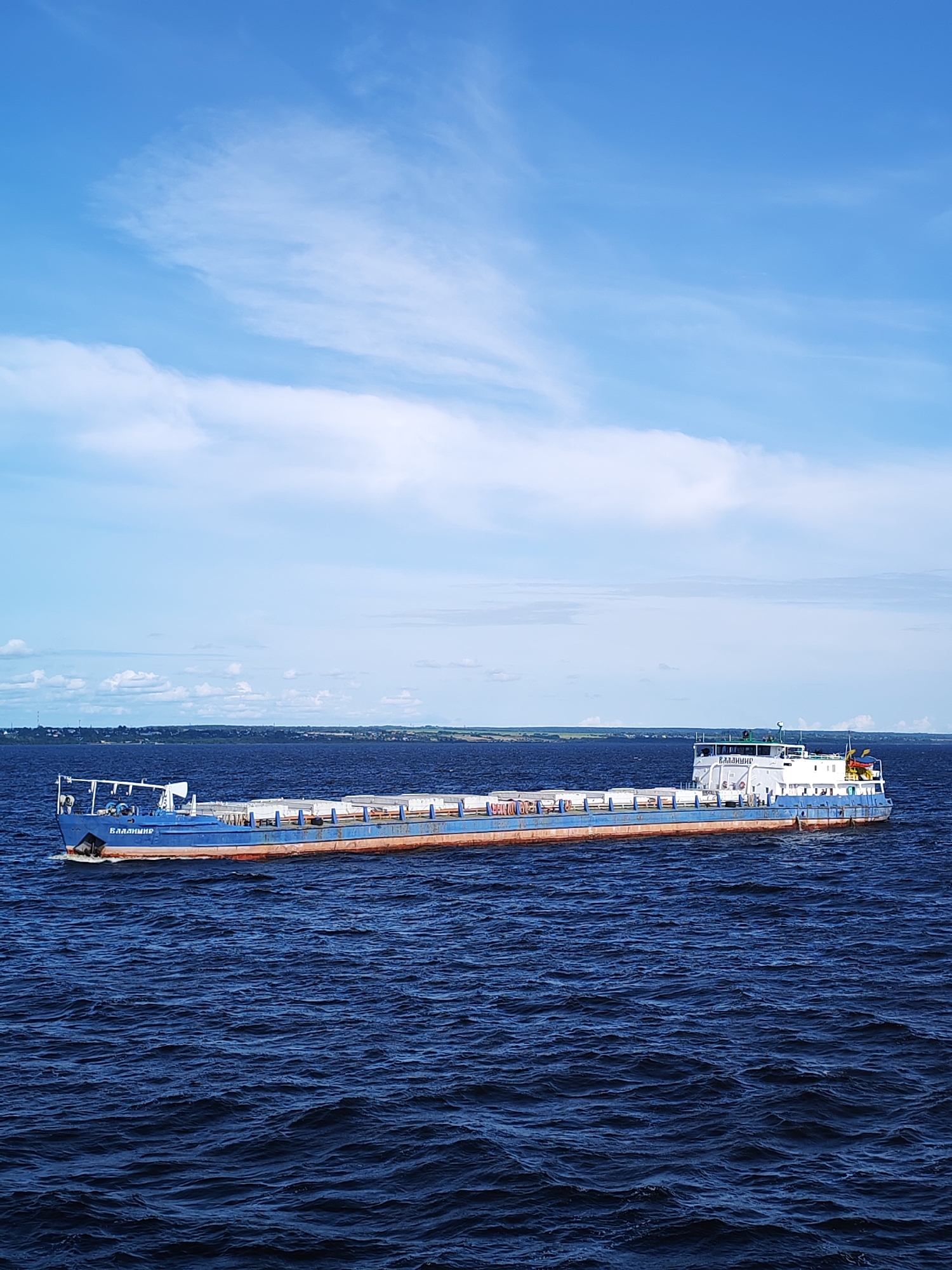 A cargo barge, identified by the name "ВЛАДИМИР" on its bow, sails on a dark blue, choppy body of water, likely a river or lake. The barge is predominantly blue with a reddish-brown waterline. It is loaded with white rectangular containers stacked along its deck. The barge is positioned in the middle ground of the image, moving from left to right. In the background, a distant, green shoreline with scattered buildings and trees is visible under a bright blue sky with scattered white clouds. The environment suggests a daytime scene under a clear sky. The location is the city of Myaksa, Russia.